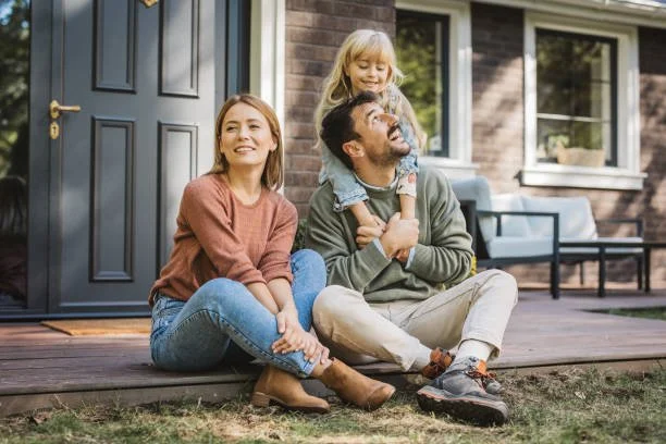 Happy family sitting on the front porch of their brick home, with a young daughter on her father's shoulders.