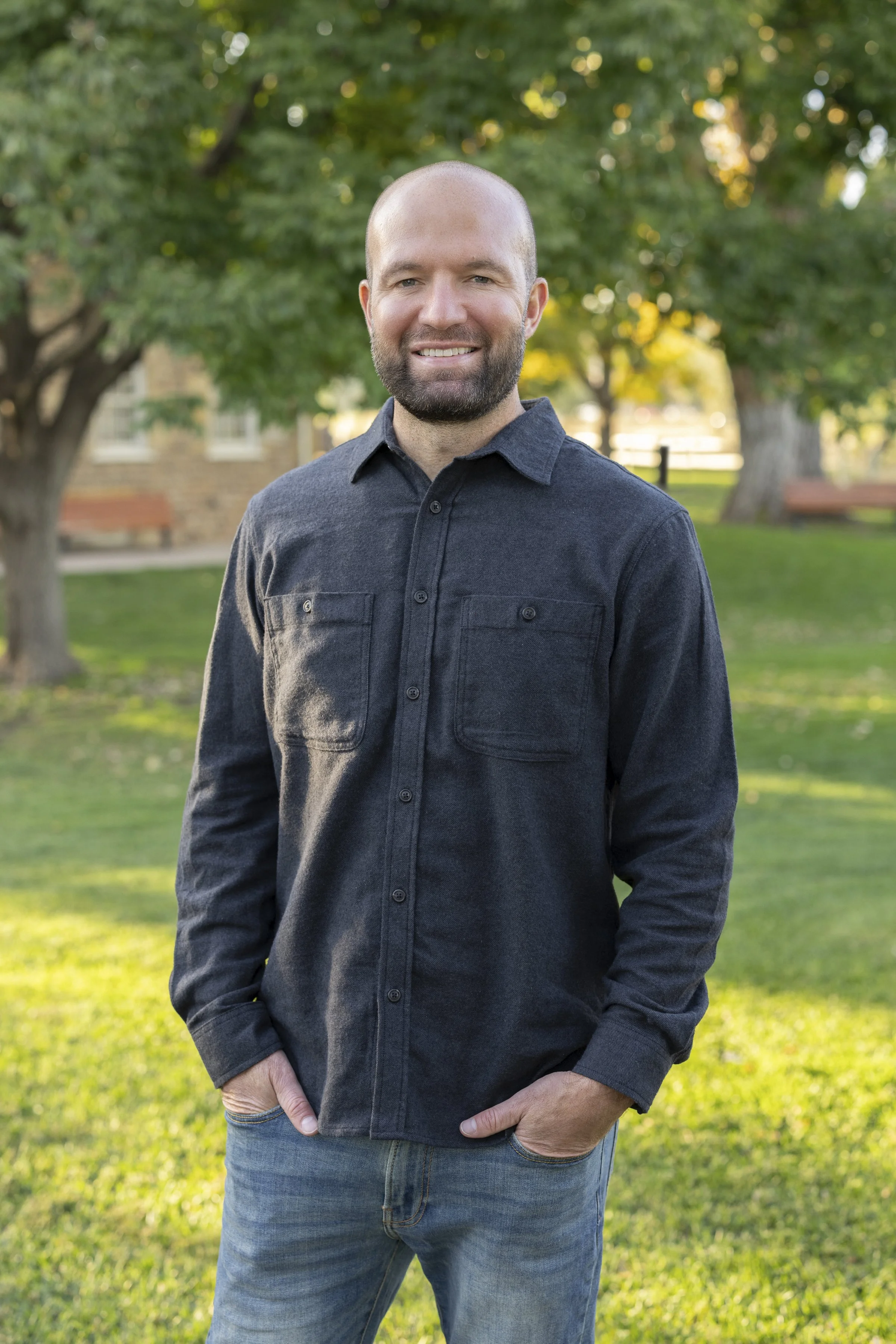Smiling bald man with a beard wearing a dark button-up shirt outdoors in a sunny park