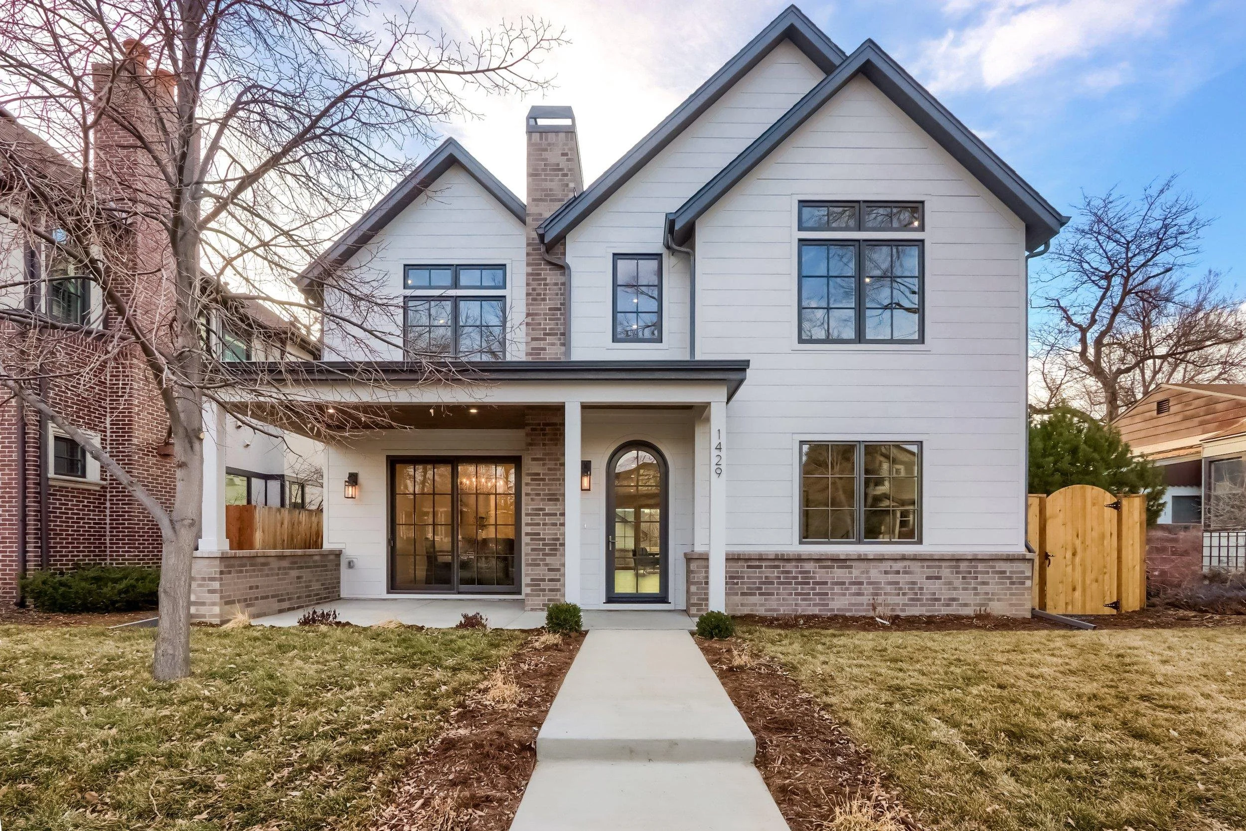 Modern two-story white-and-brick house with gabled roof, covered porch, large windows, front walkway, and leafless trees.