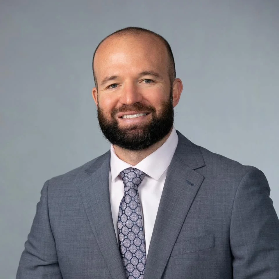 Professional headshot of a smiling bald man with a beard wearing a gray suit, white shirt, and patterned tie against a gray background