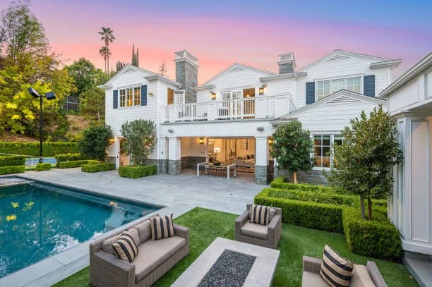 Luxury white two-story home with poolside patio, manicured hedges, outdoor seating, and sunset sky