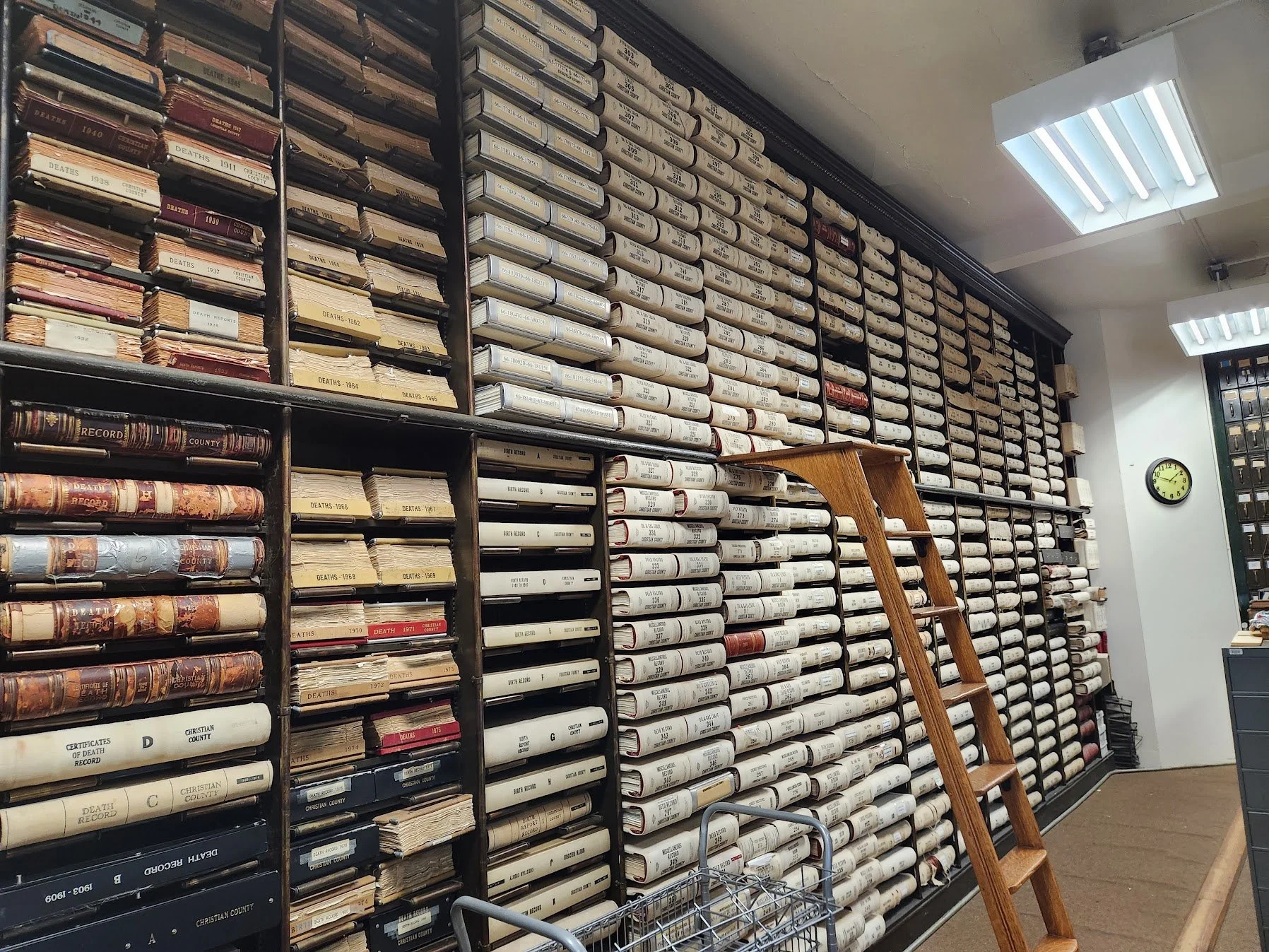 Historic county death records and bound volumes lining archive shelves, with a wooden ladder and fluorescent lights