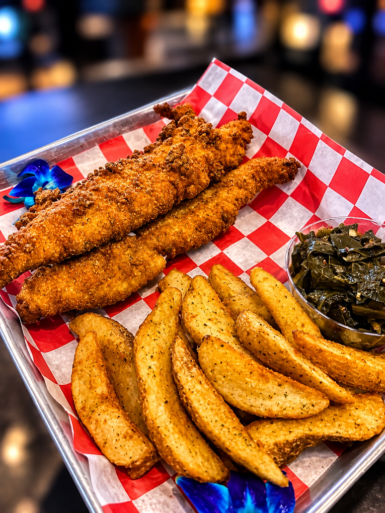 Basket of crispy breaded fish fillets with seasoned potato wedges and collard greens on red-and-white checkered paper