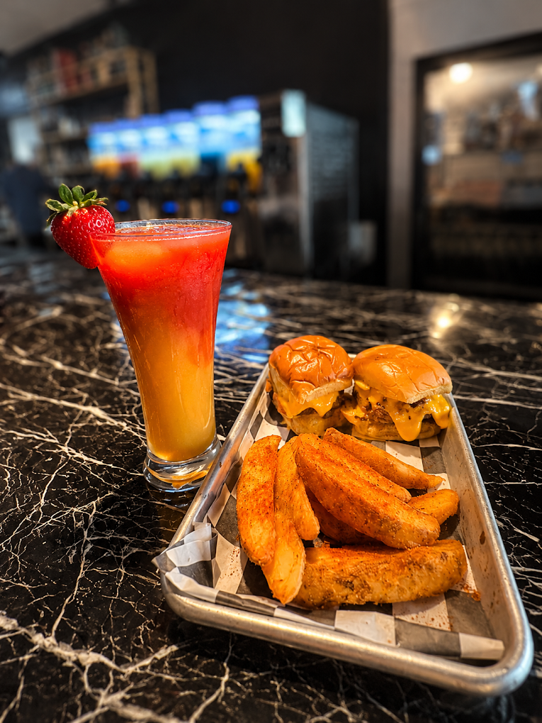 Cheeseburger sliders, seasoned potato wedges, and a strawberry-garnished orange-red cocktail on a restaurant table