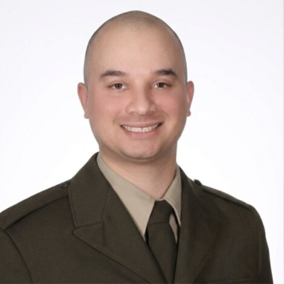 Smiling bald man in a brown military-style uniform and tie against a white background