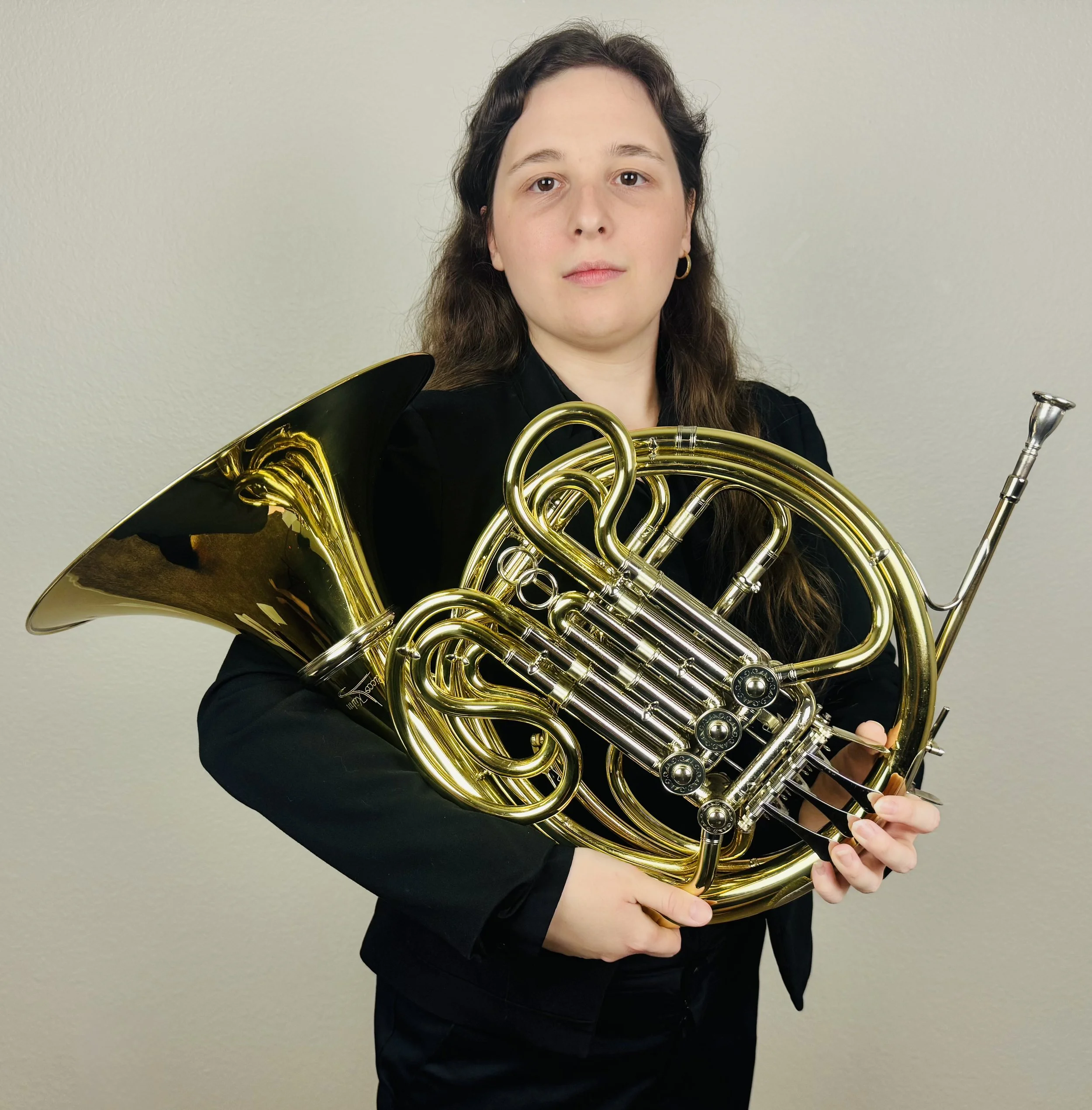 Musician in black holding a polished brass French horn against a neutral wall