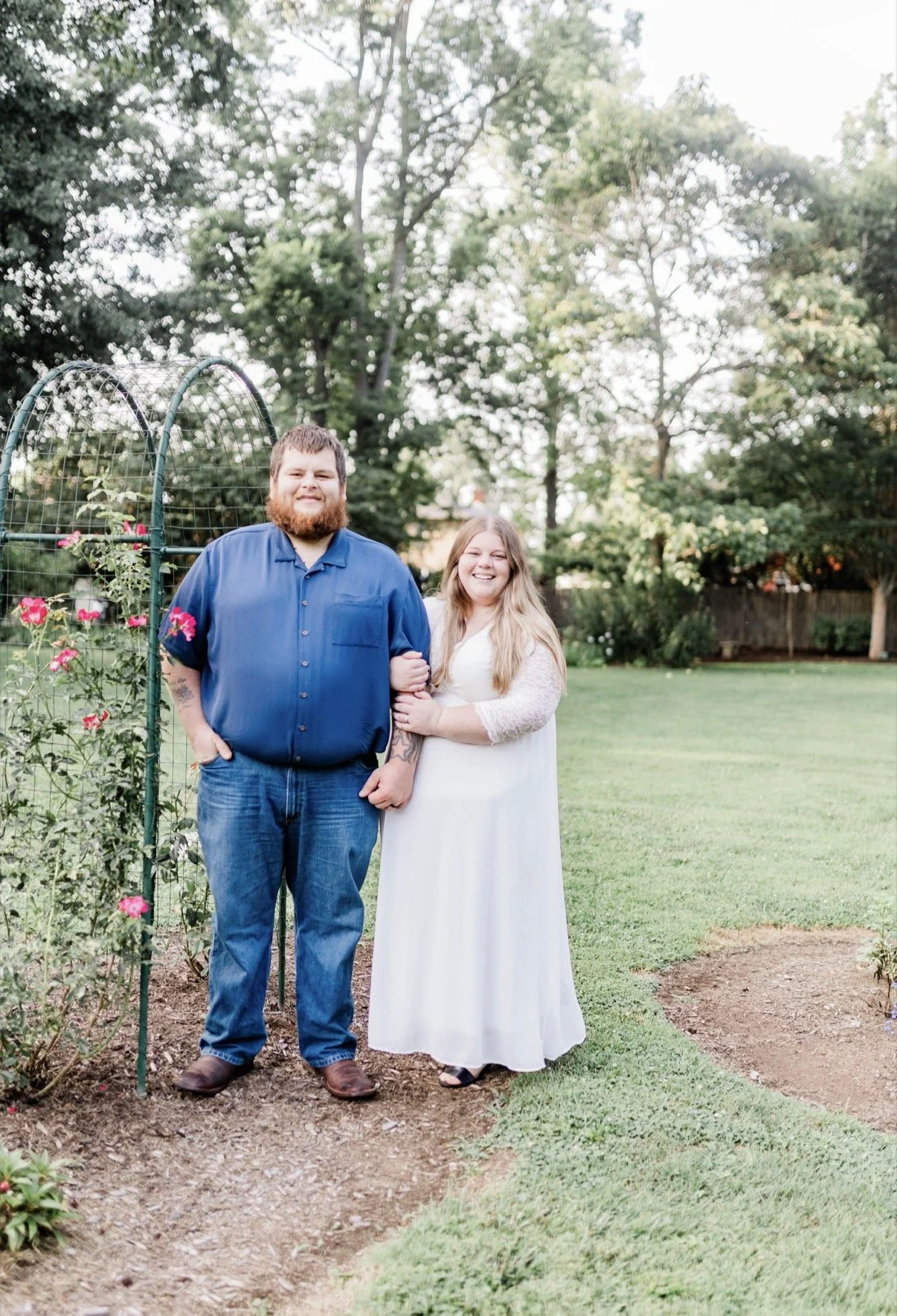Smiling couple standing together beside a rose-covered garden trellis in a backyard.
