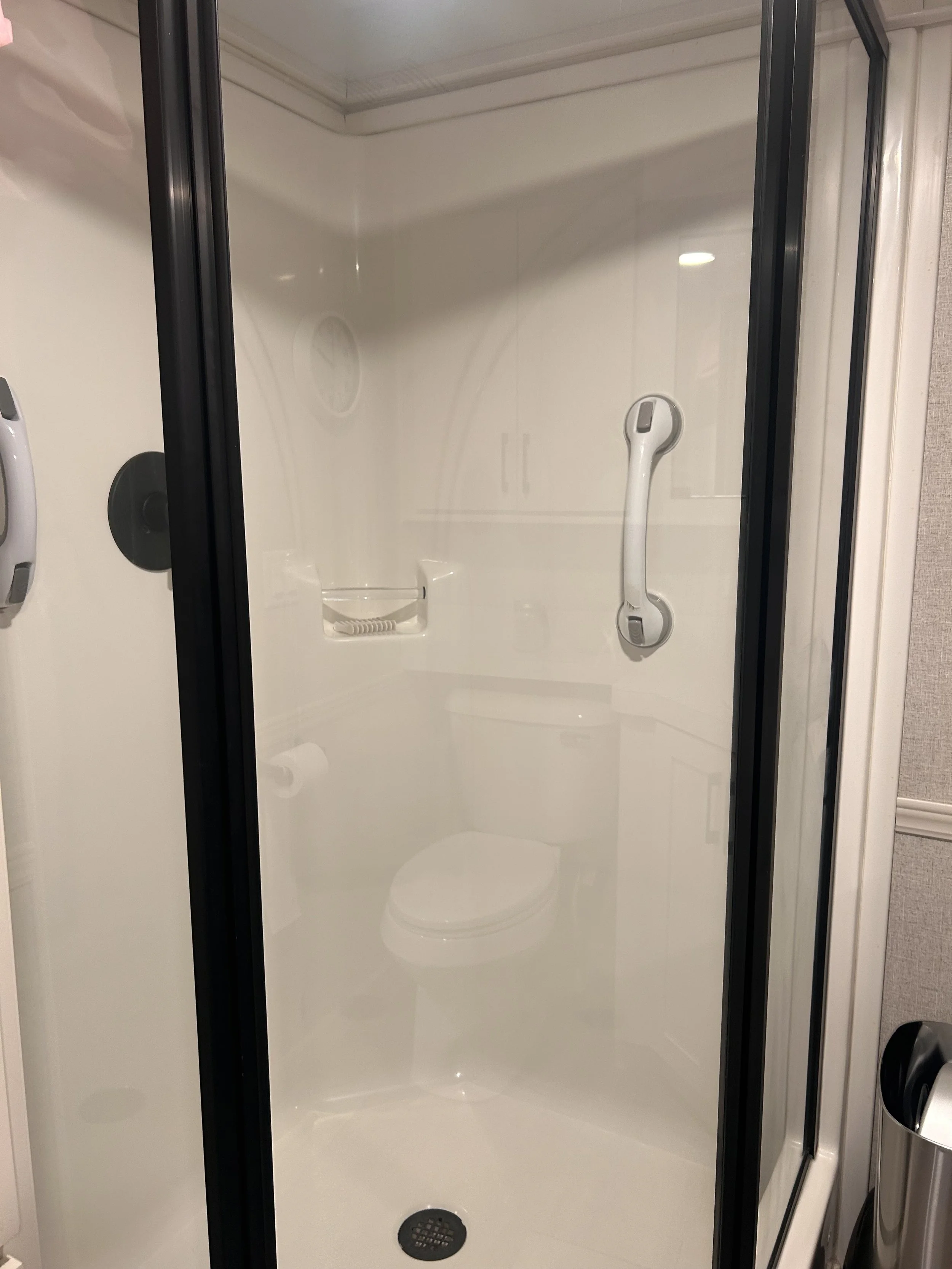 White fiberglass RV shower stall with black-framed glass doors, built-in seat, grab handle, and drain