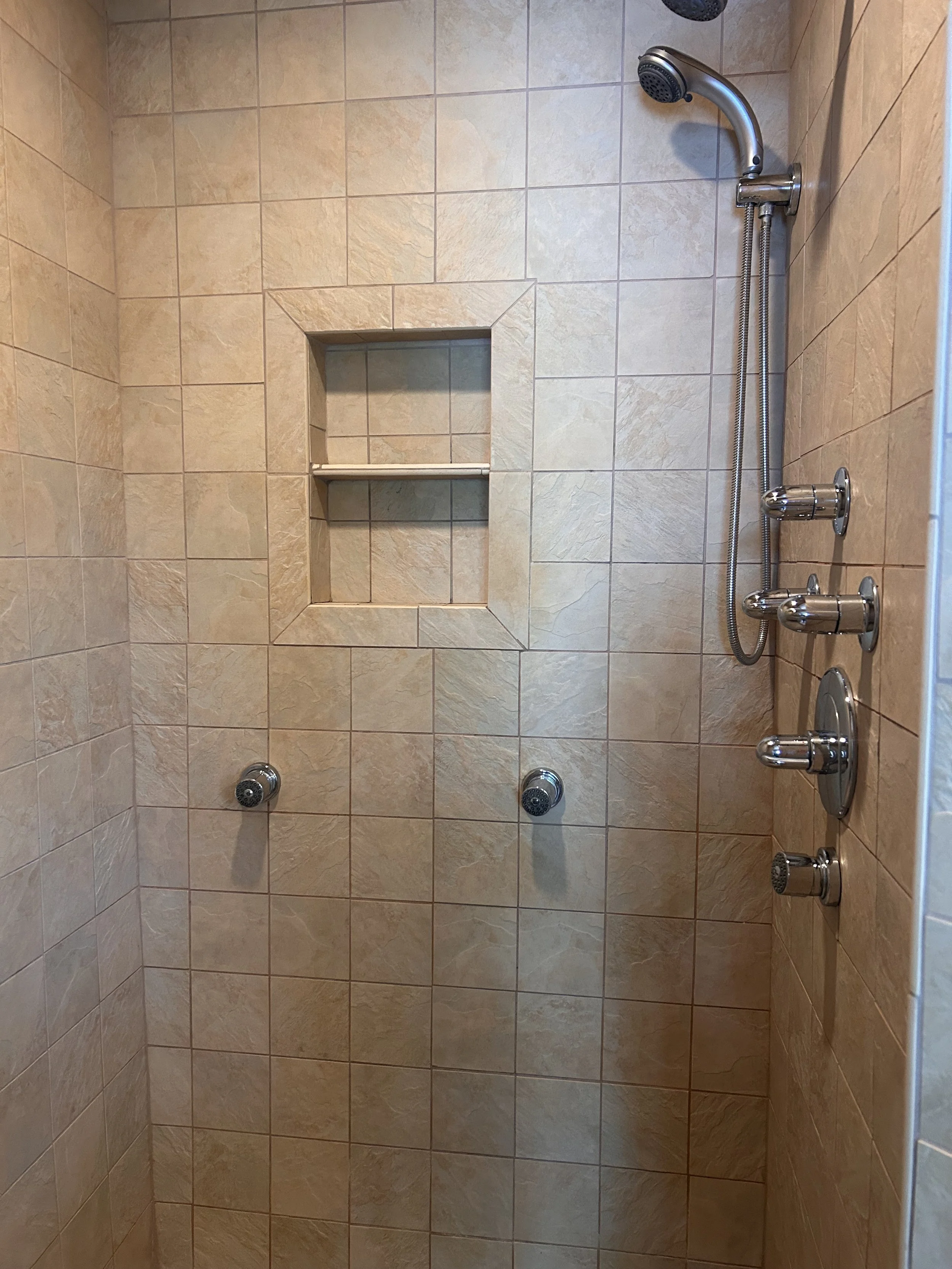 Tiled shower stall with multiple chrome showerheads, handheld shower hose, body jets, and recessed shelf