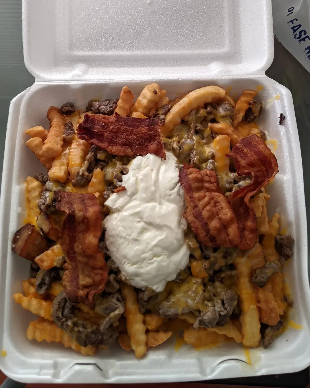 Loaded cheese fries topped with ground beef, bacon, and sour cream in a foam takeout container