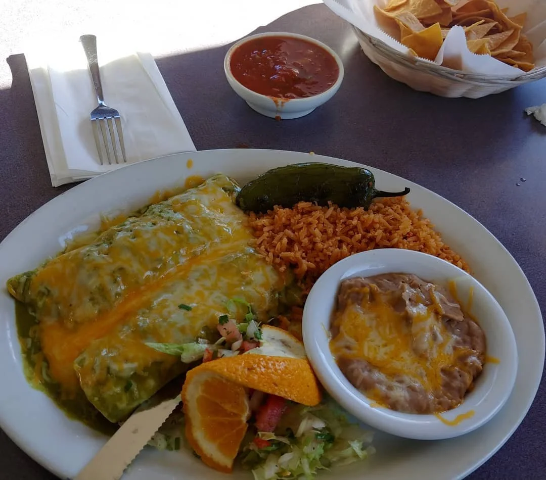Mexican restaurant plate with a cheese-covered green chile enchilada, Spanish rice, refried beans, salad, and orange slices, served with salsa and tortilla chips