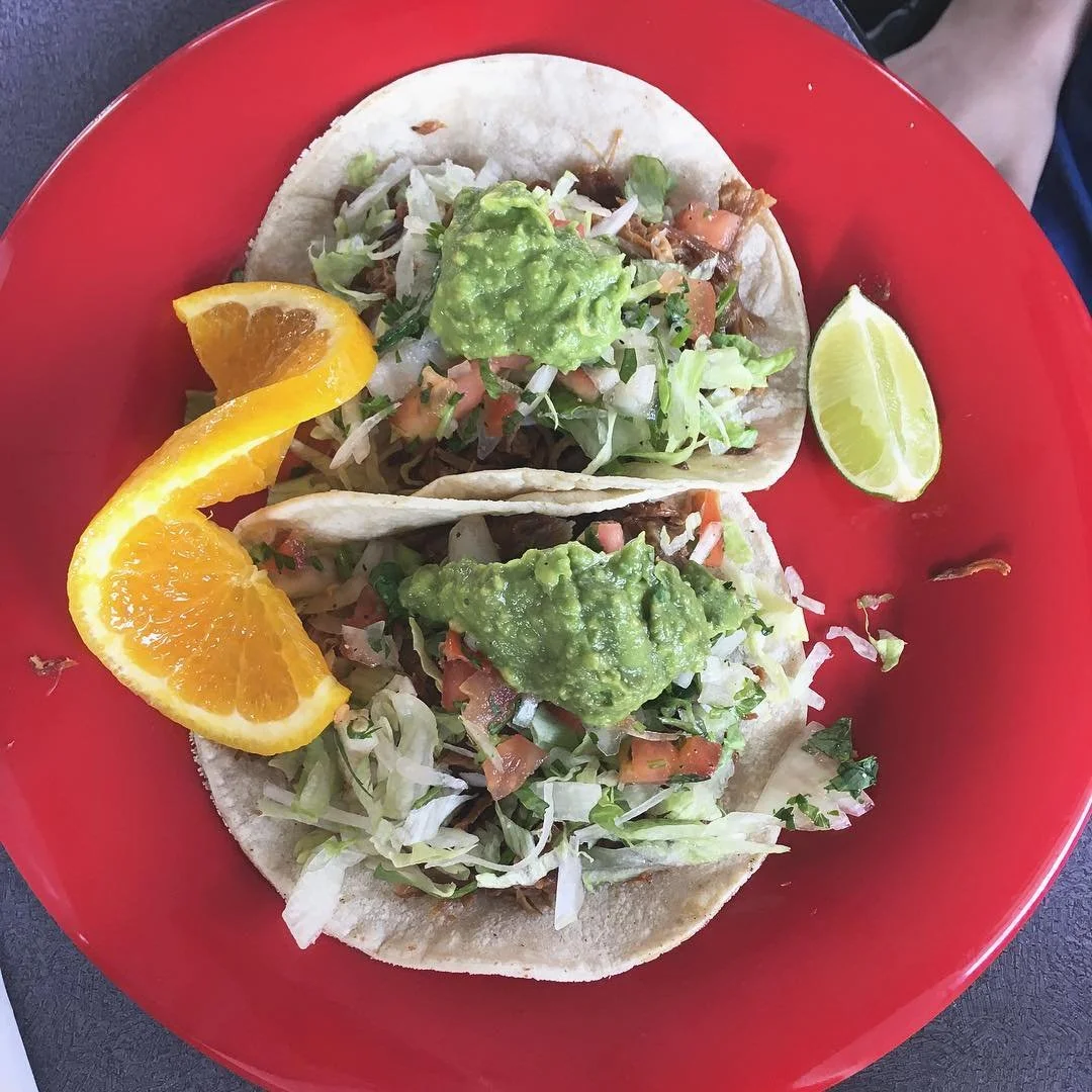 Two soft tacos topped with guacamole, lettuce, pico de gallo, and cilantro on a red plate with orange and lime wedges