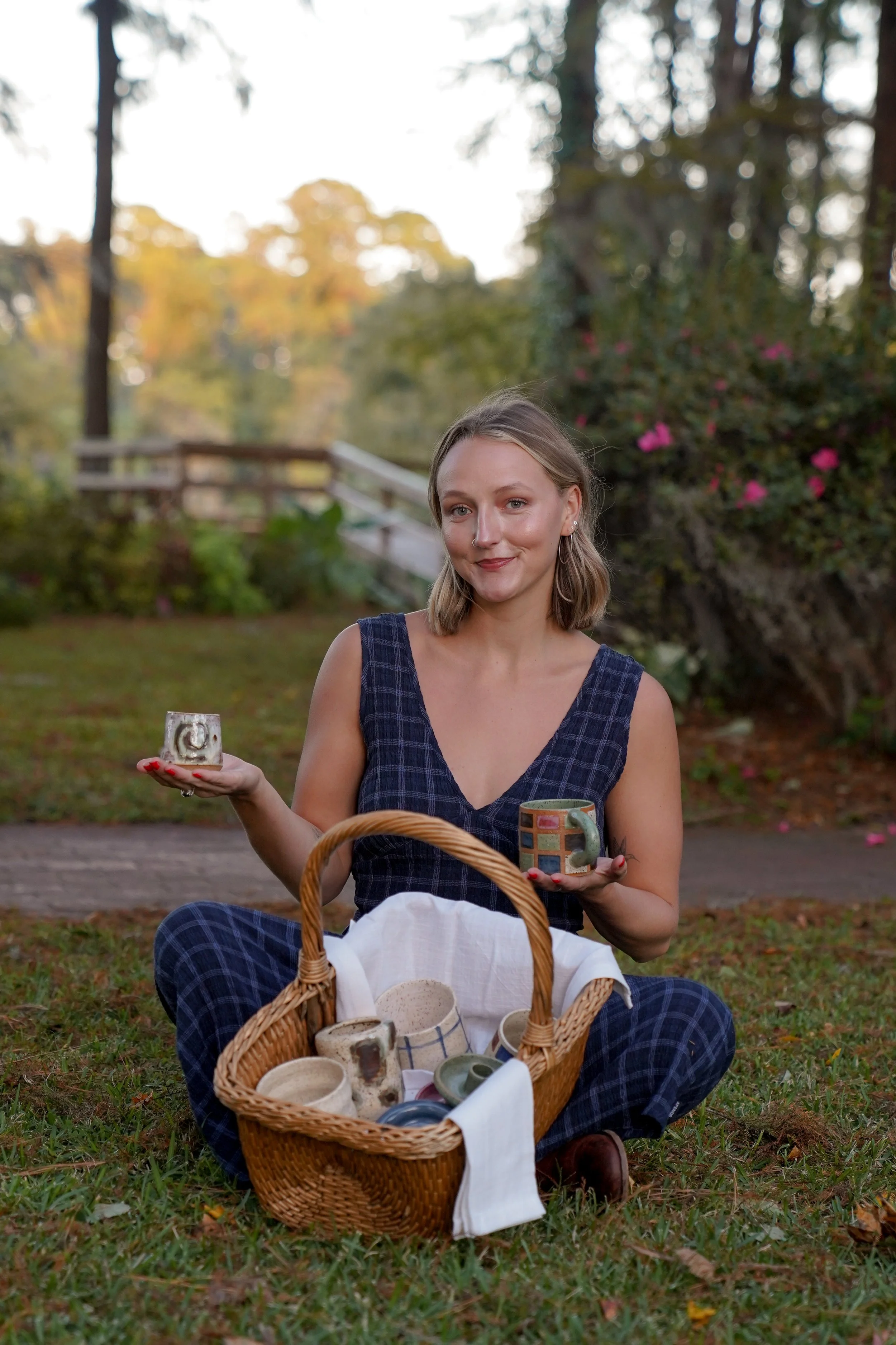 Woman in a garden holding handmade ceramic cups beside a wicker picnic basket filled with pottery.
