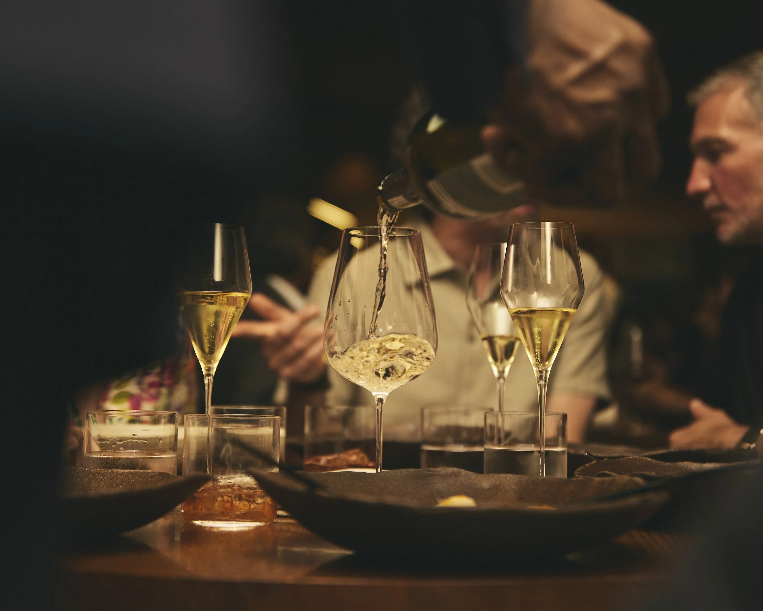 Bartender pouring sparkling wine into a glass at a dimly lit restaurant bar surrounded by wine glasses