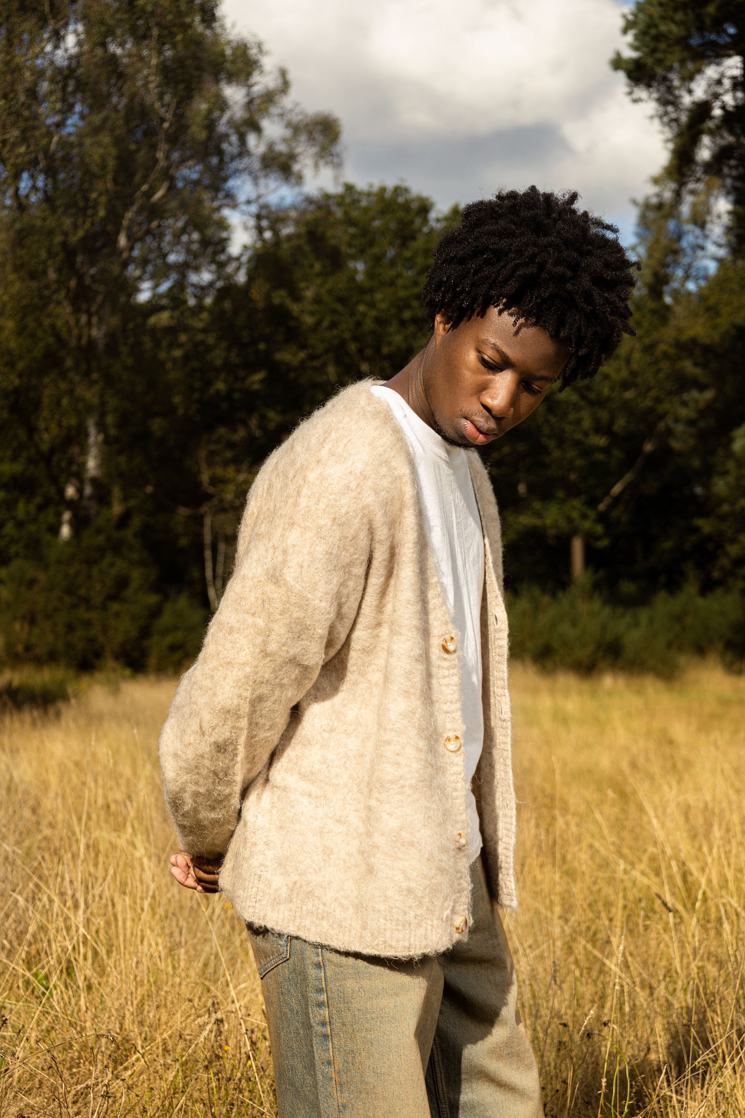 Outdoor fashion portrait of a male model wearing a cream cardigan and neutral denim in a sunlit field, photographed by Ade West.