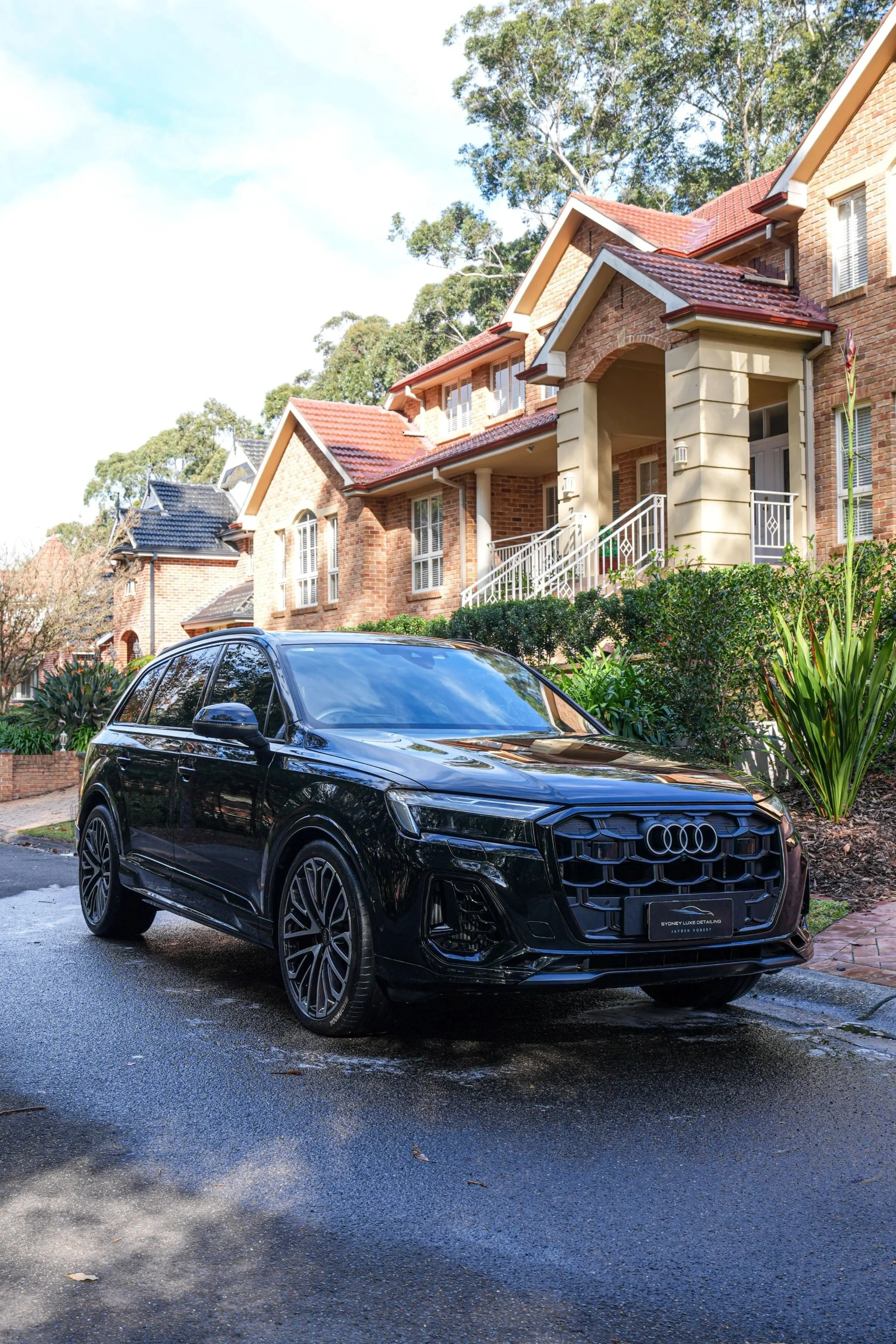 Black Audi SUV parked in front of a brick suburban home with red tile roofs and lush landscaping.