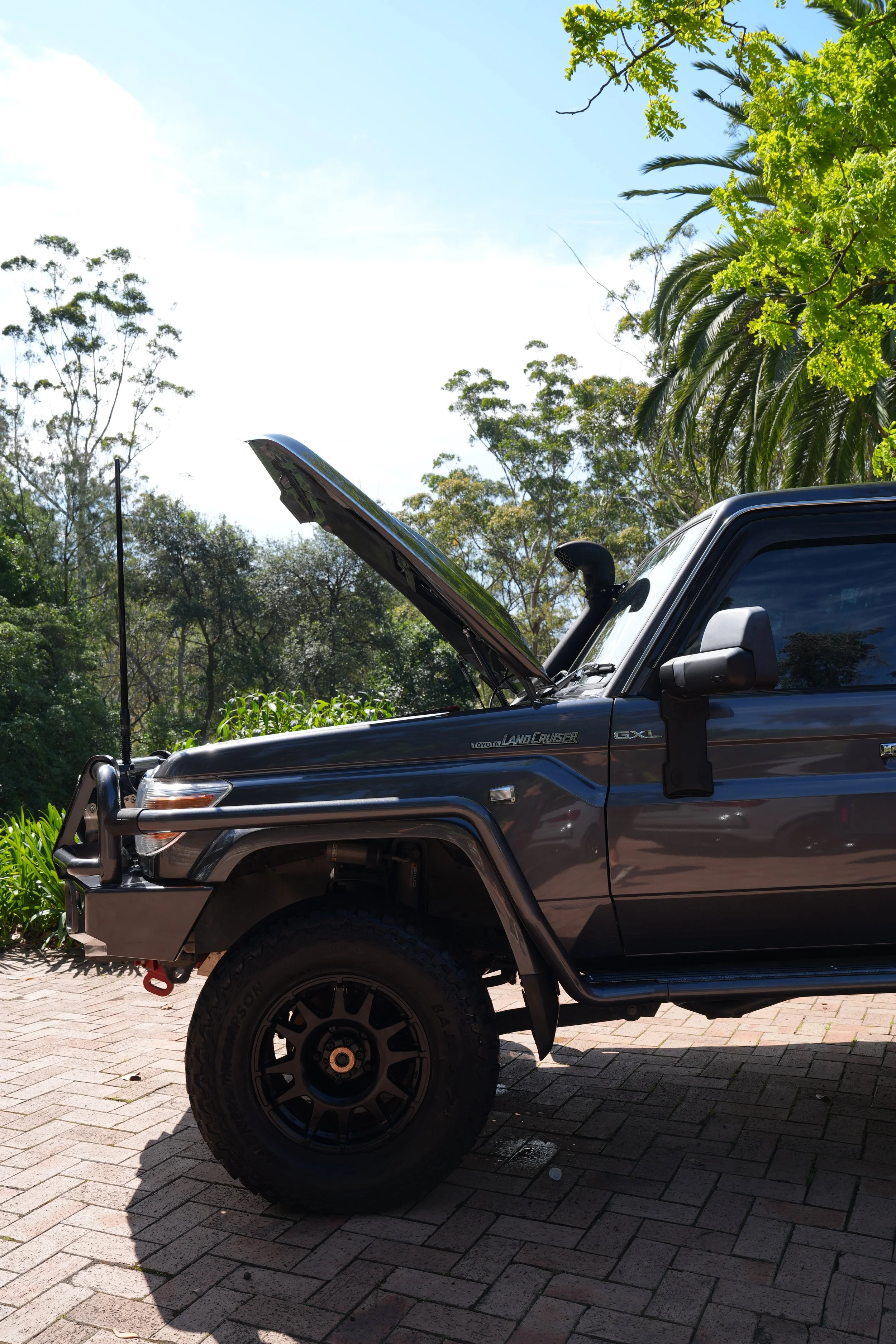 Dark gray Toyota Land Cruiser GXL pickup truck with raised hood, bull bar, snorkel, and off-road tires parked outdoors among trees.