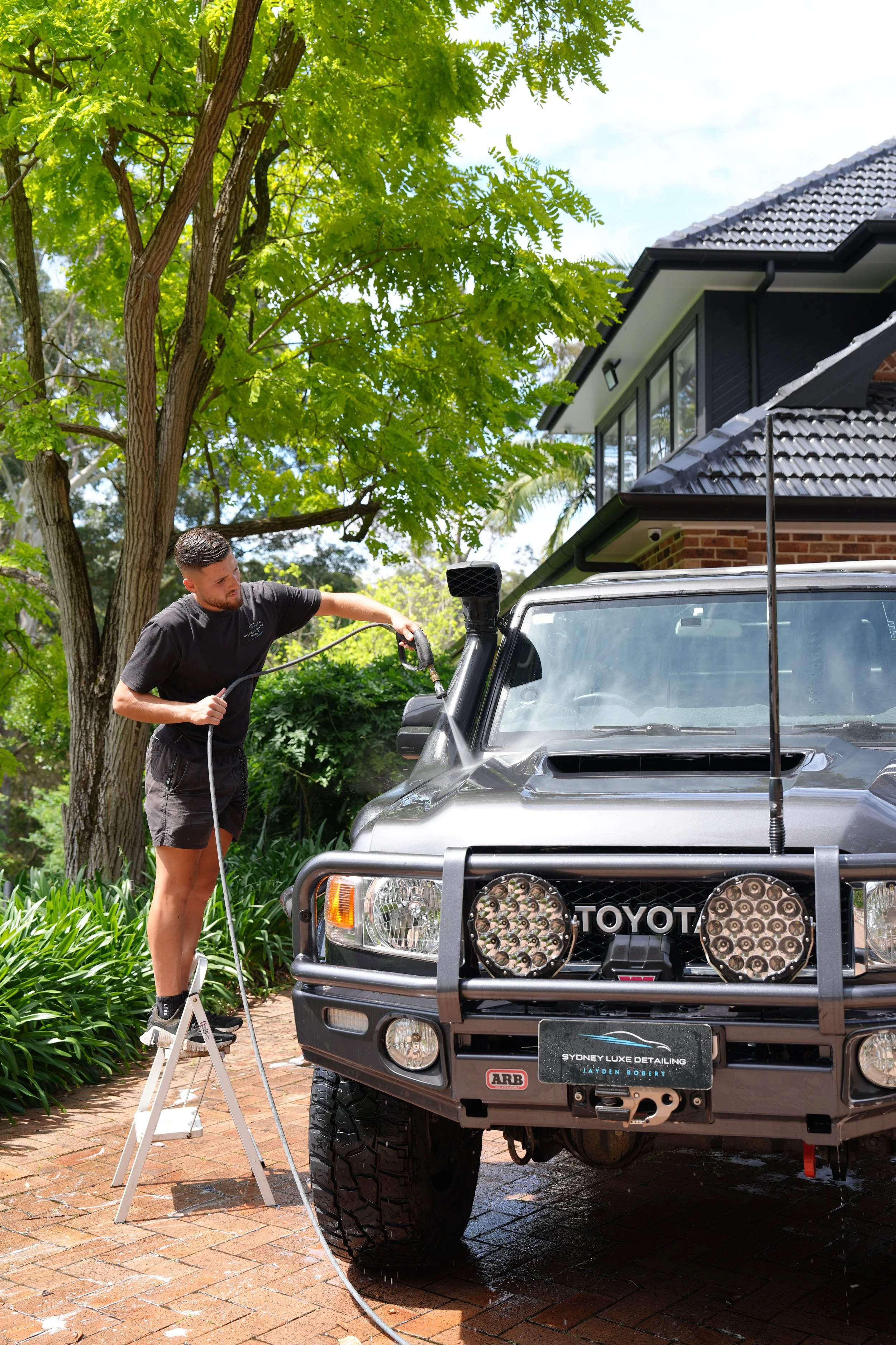 Detailer pressure-washing the front of a Toyota 4WD SUV in a residential driveway.