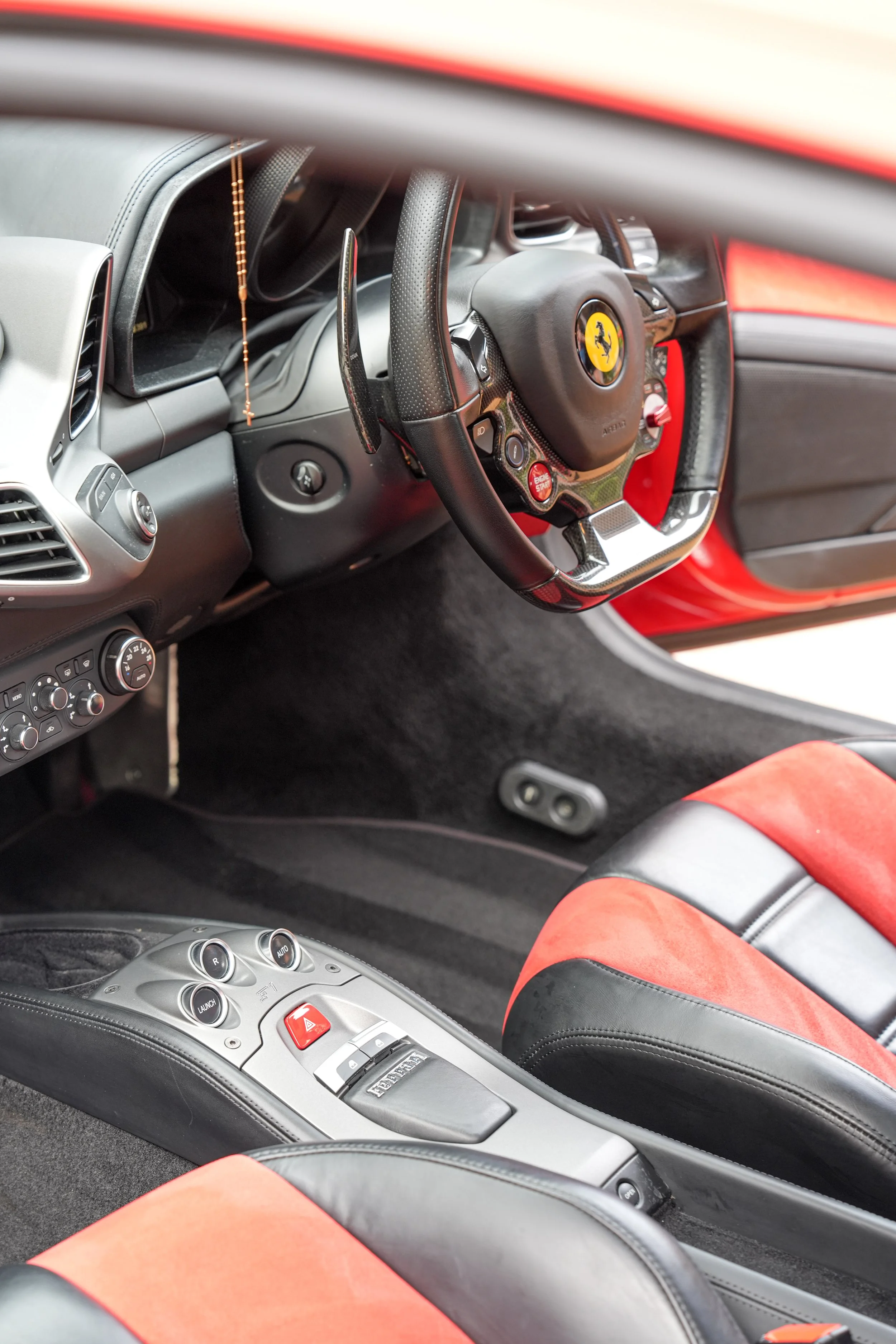 Interior of a Ferrari sports car showing the steering wheel, carbon-fiber dashboard, red start button, center console, and red-and-black leather seats.