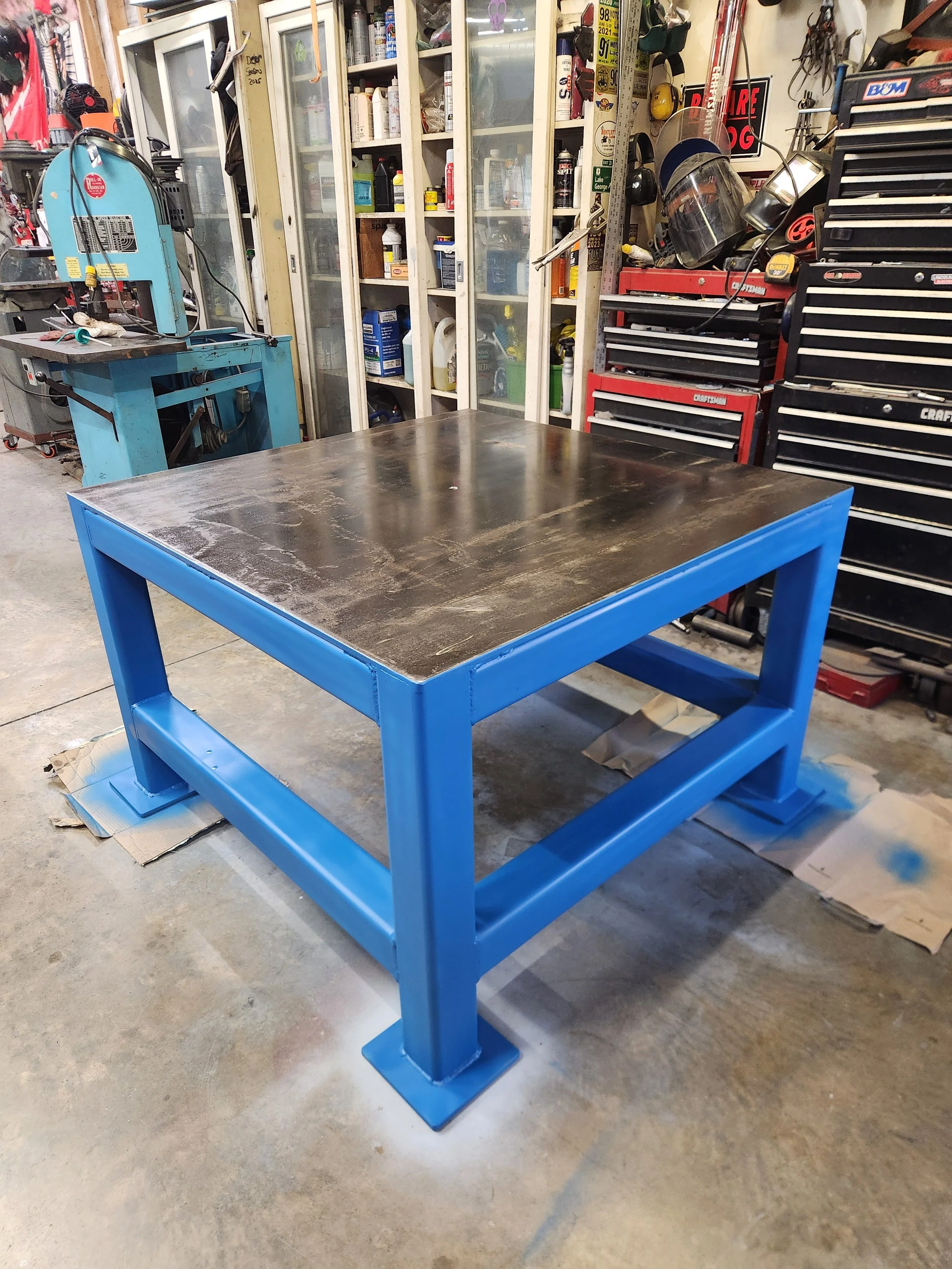 Blue metal workbench with a steel tabletop in a cluttered auto repair garage, surrounded by tool chests, cabinets, and equipment.