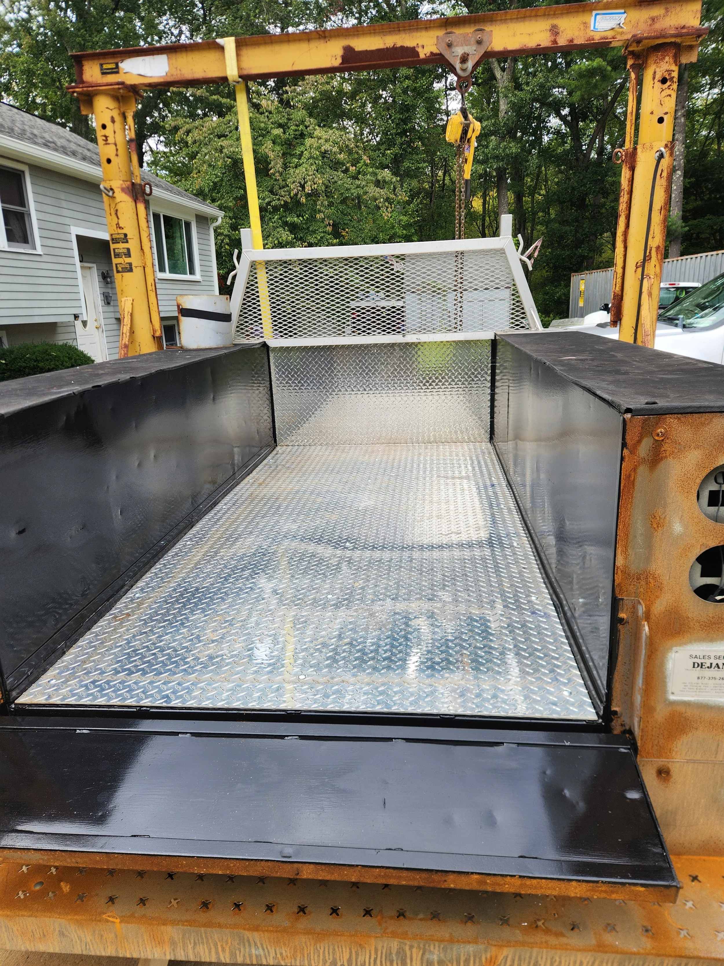 Rusty yellow truck-mounted crane lifting a white metal landscaping trailer beside a house and wooded yard
