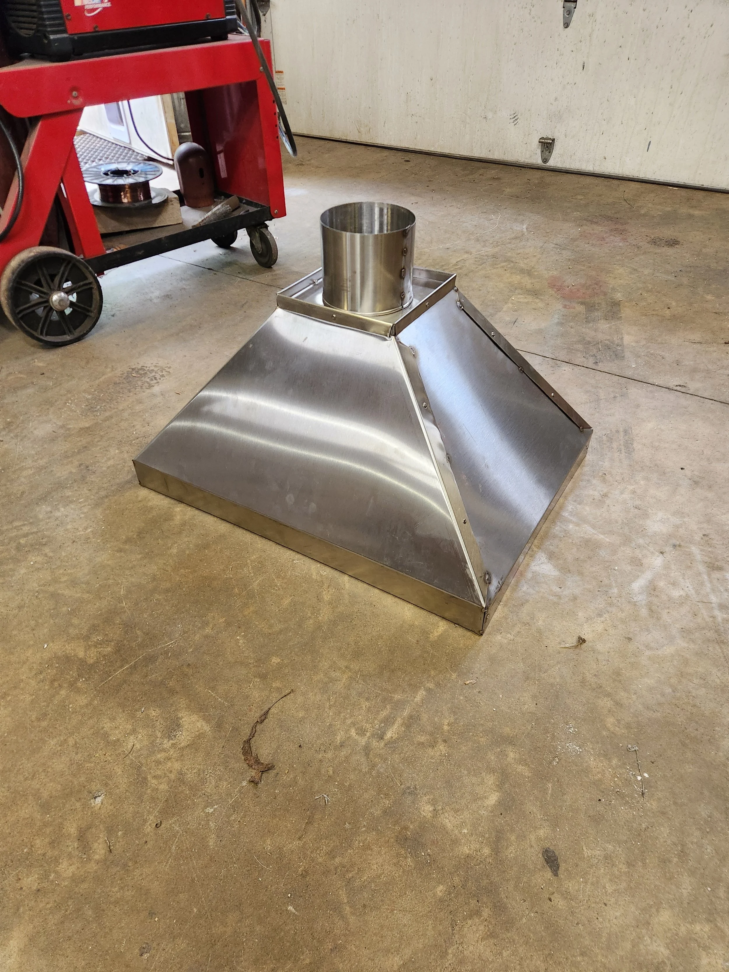 Stainless steel industrial hopper beside a red wheeled fabrication cart in a workshop