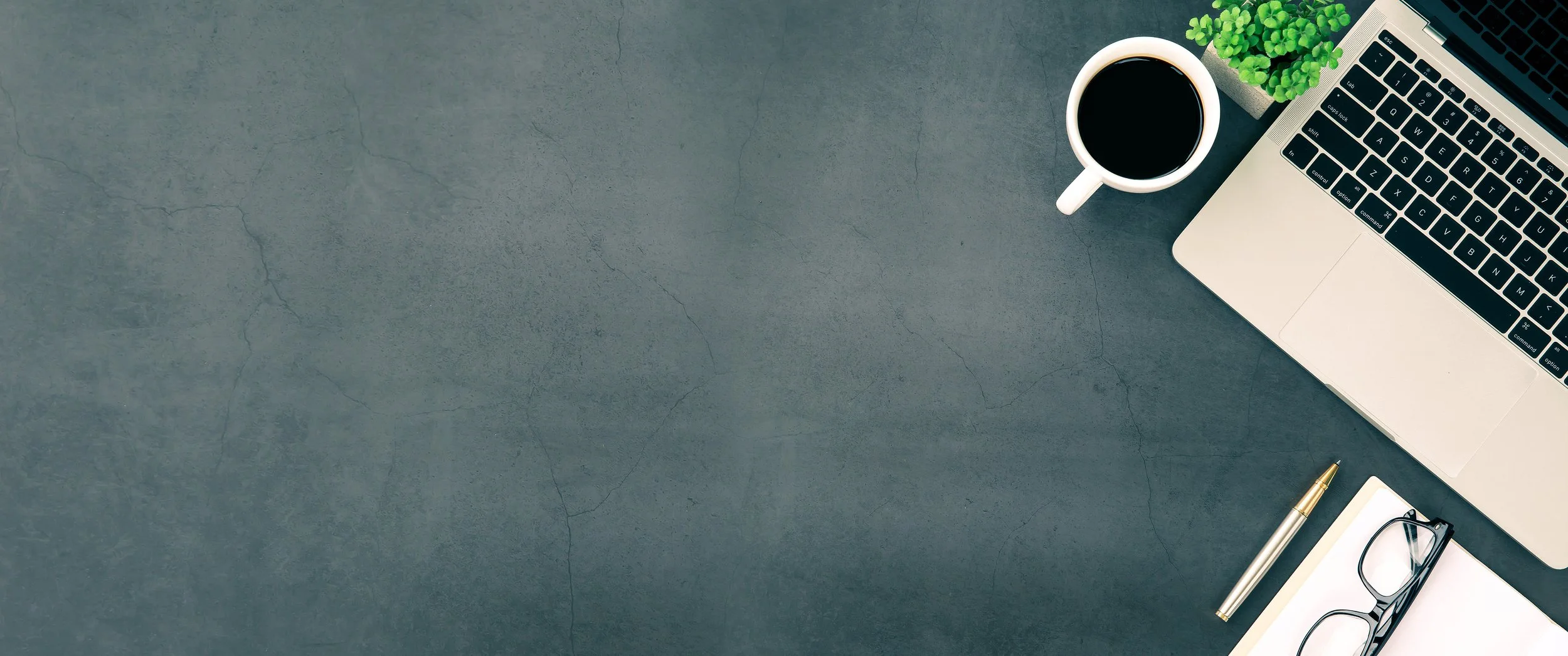 Top-down view of a laptop, coffee cup, plant, pen, notebook, and eyeglasses on a dark desk with copy space