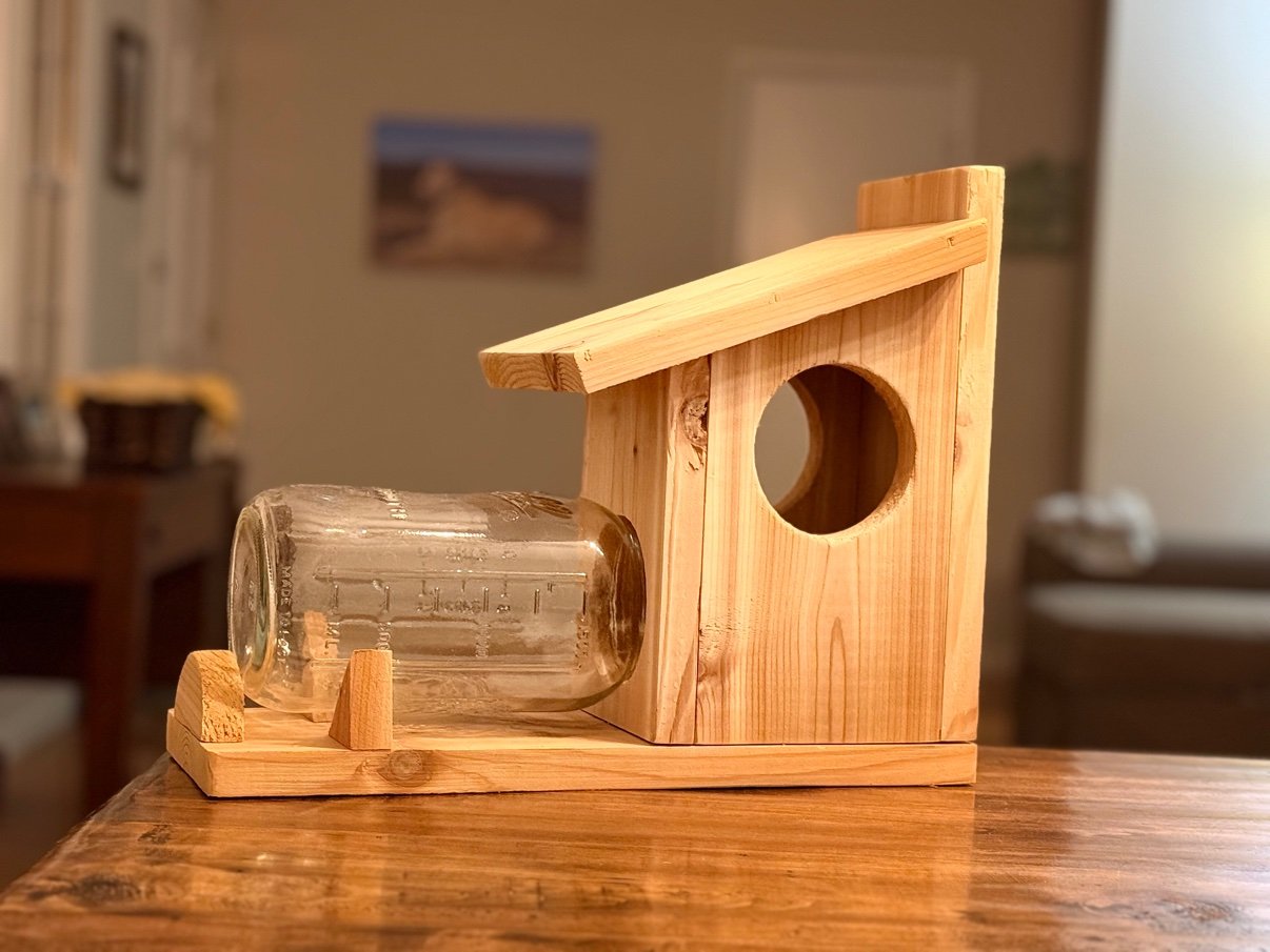 Side view of Cedar Squirrel Feeder indoors, with slanted roof, large round opening, and empty clear glass mason jar lying horizontally on the platform