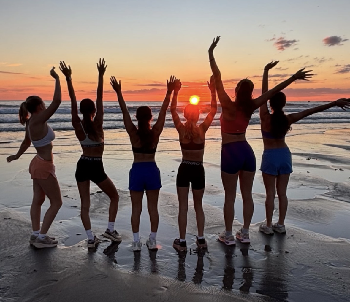 Group of women raising their arms on a beach at sunset, with ocean waves and reflections on the wet sand