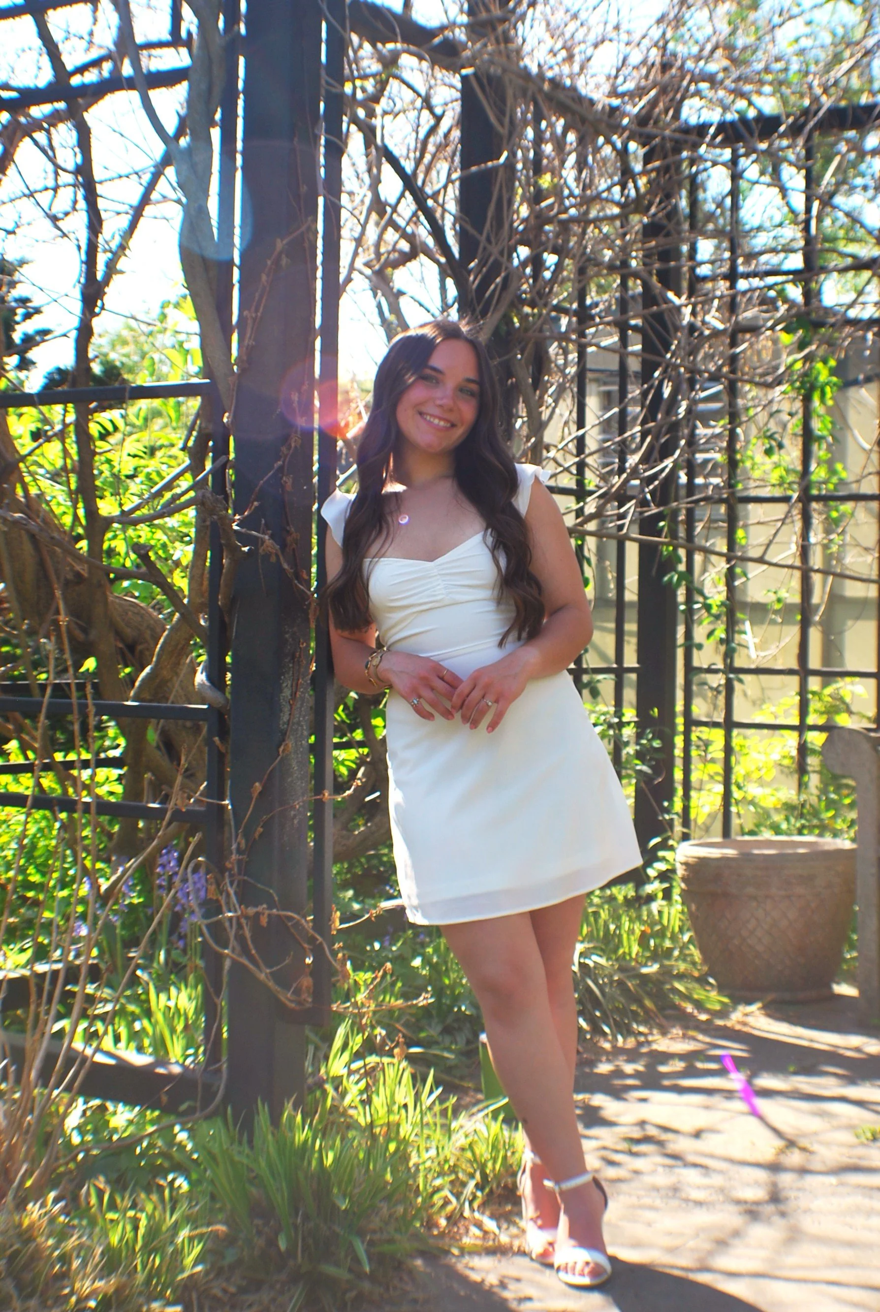 Smiling woman in a white dress posing outdoors among trees and greenery.