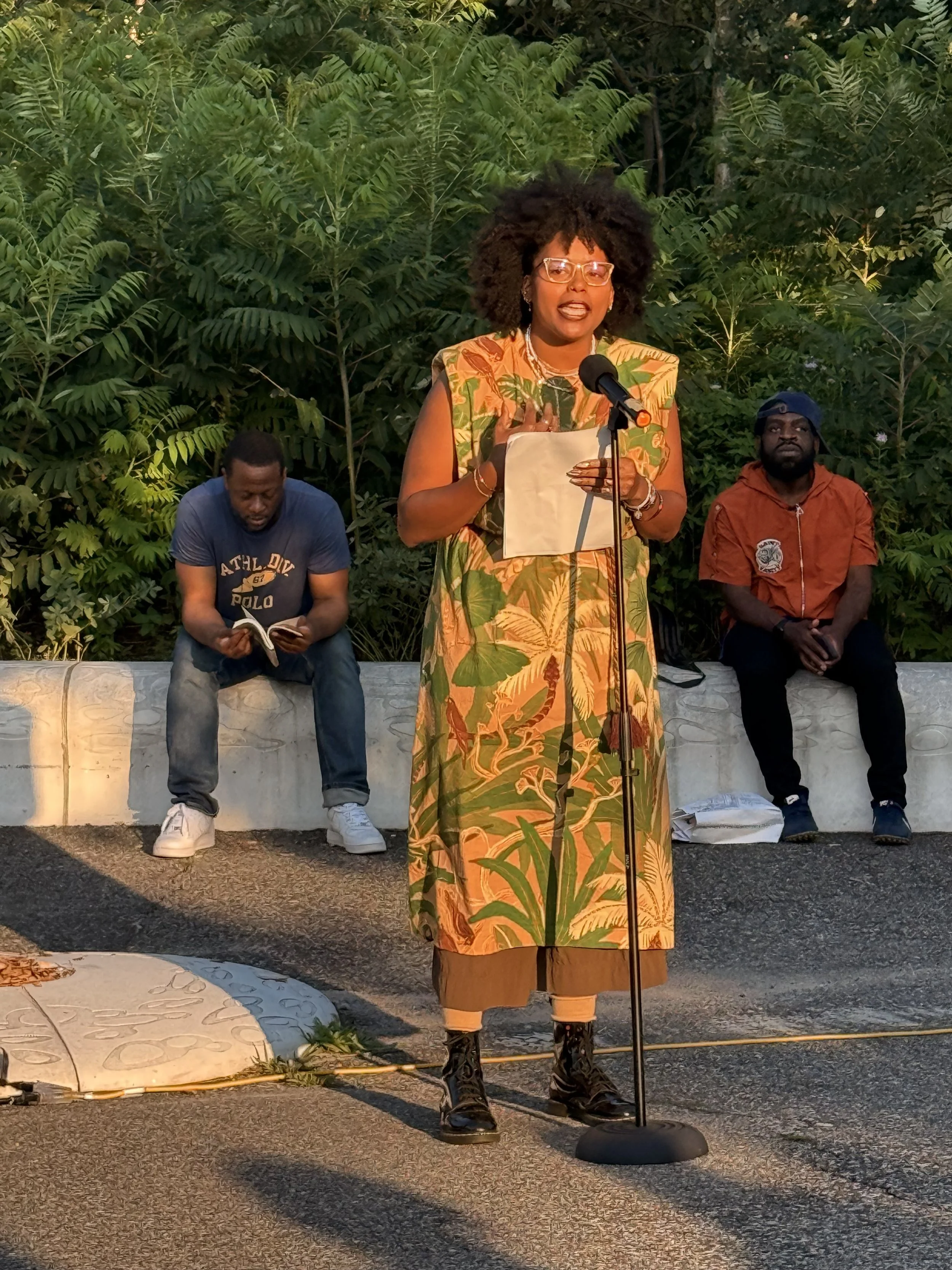 Woman in a colorful patterned dress speaking into a microphone outdoors, with two seated men and lush foliage behind her