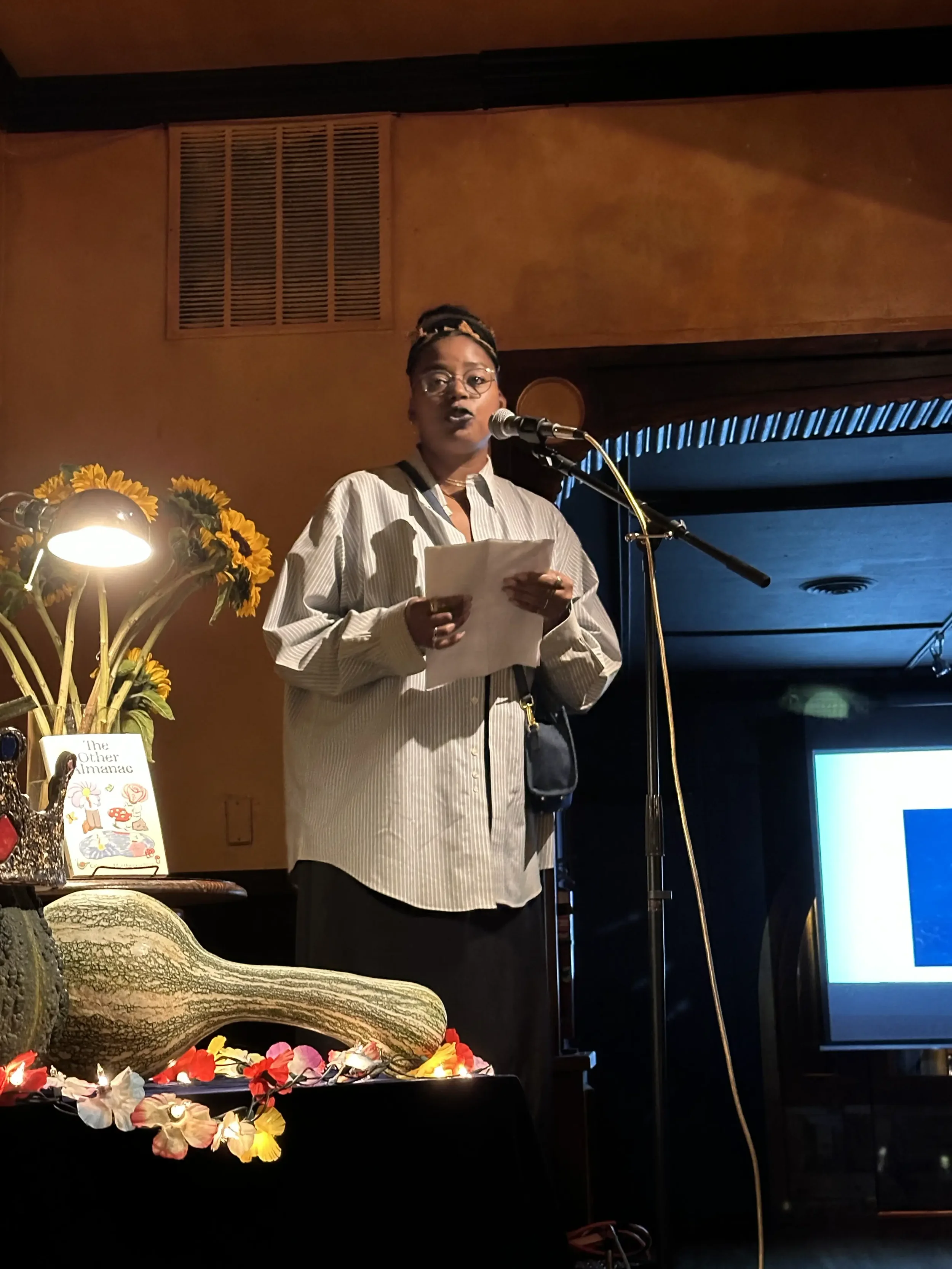 Woman reading from papers at a microphone onstage, with sunflowers, a book, and decorative gourds nearby.