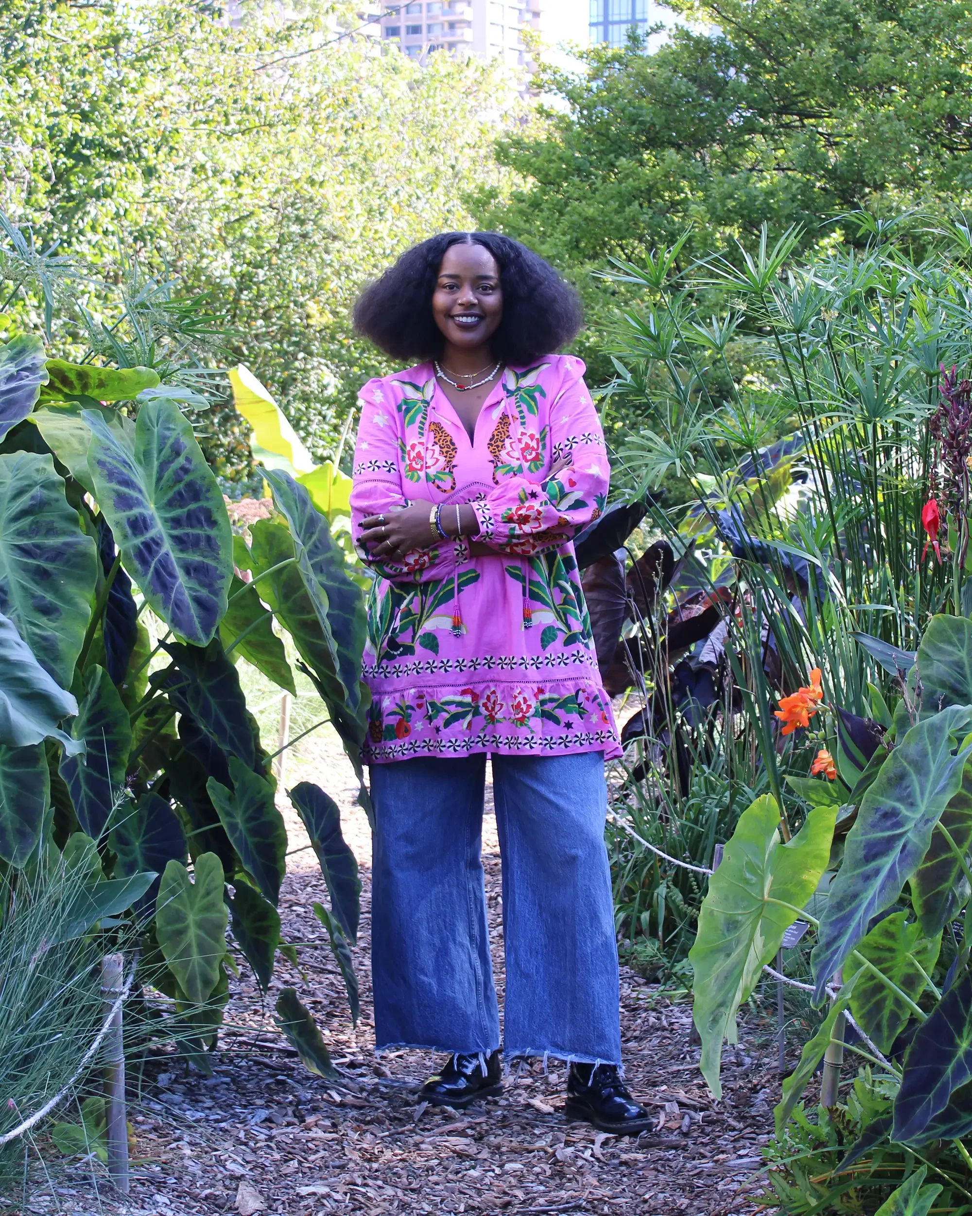 Smiling woman in a colorful pink patterned blouse standing among lush tropical plants in a botanical garden