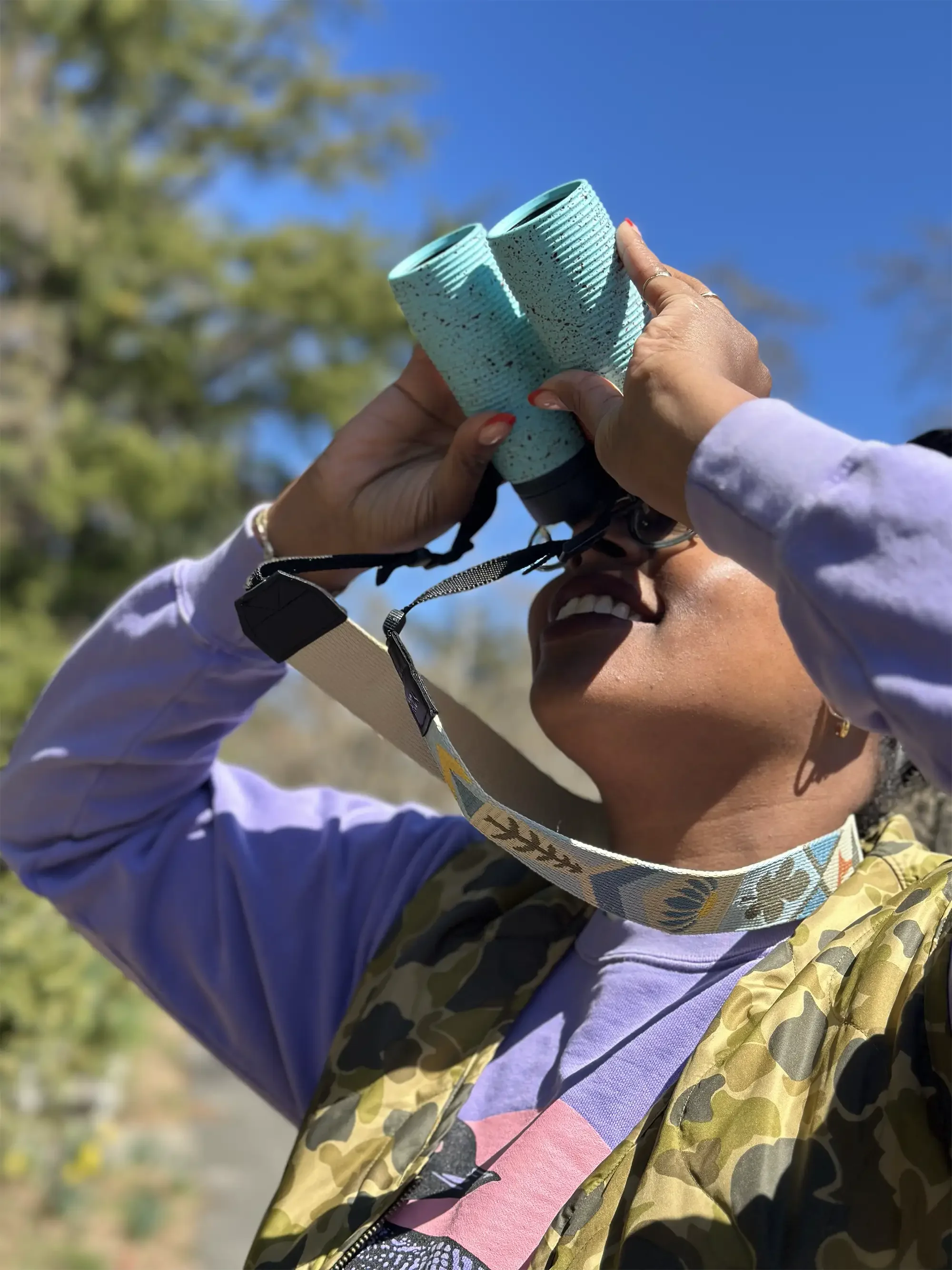 Person smiling while holding turquoise binoculars outdoors against a blue sky
