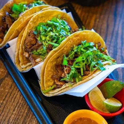 Three street-style beef tacos topped with chopped cilantro and onions, served with lime wedges and salsa.