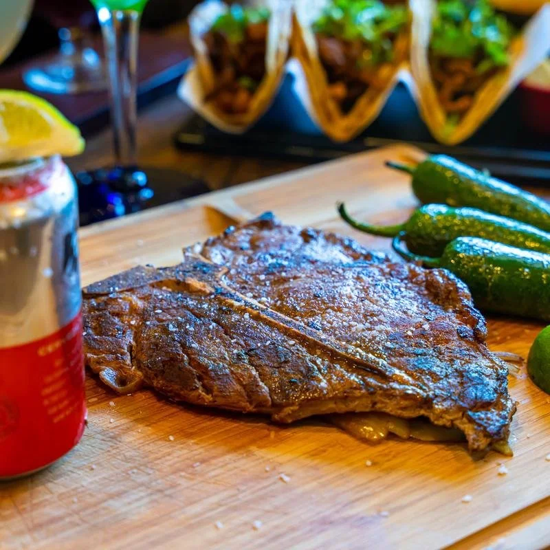 Grilled steak with roasted jalapeños on a wooden cutting board, with tacos and a lime drink in the background
