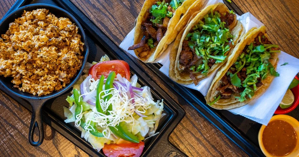 Mexican meal with four cilantro-topped tacos, scrambled eggs, salad, lime wedges, and salsa on a wooden table