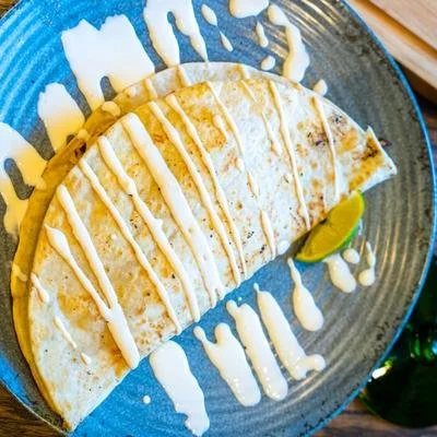Crispy folded quesadilla drizzled with creamy sauce on a blue ceramic plate, served with lime wedges.