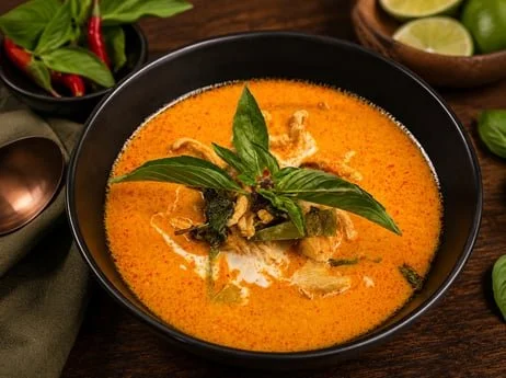 Bowl of creamy orange Thai-style curry topped with basil, served with lime wedges and red chilies