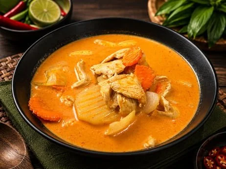 Bowl of creamy orange Thai curry with sliced chicken, vegetables, red chili, lime, and fresh herbs