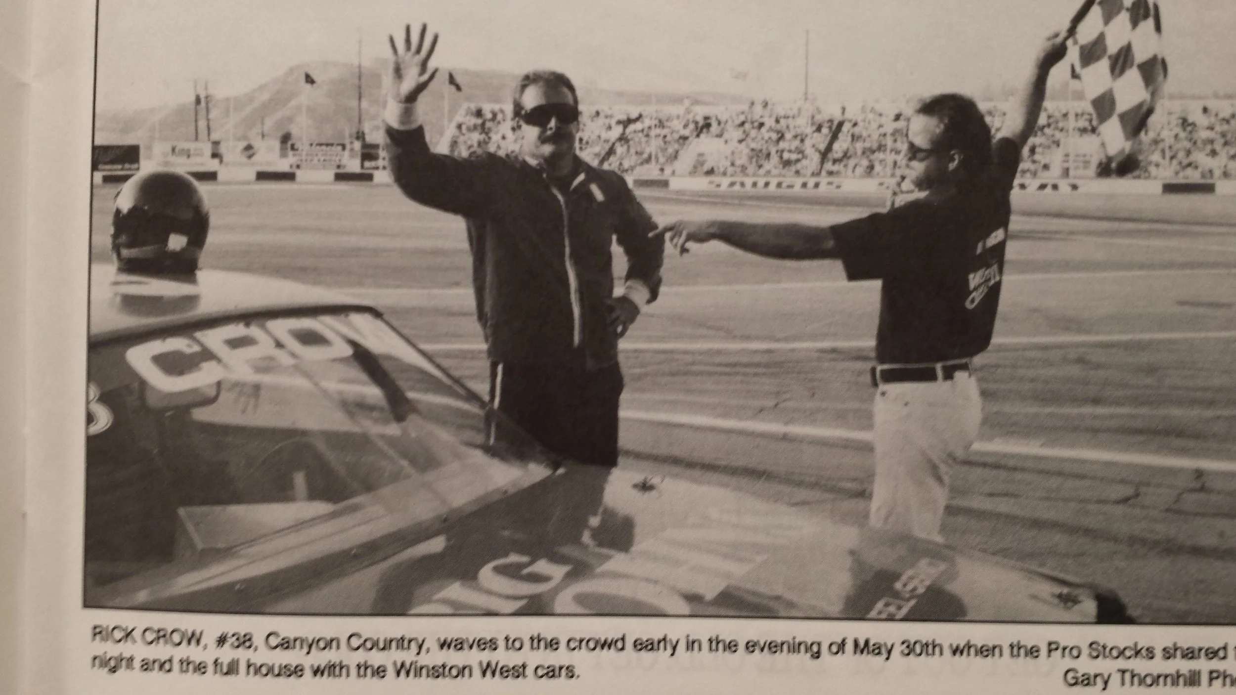 Black-and-white photo of NASCAR driver Rick Crow waving from a race car beside a crew member holding a checkered flag in Victory Lane.