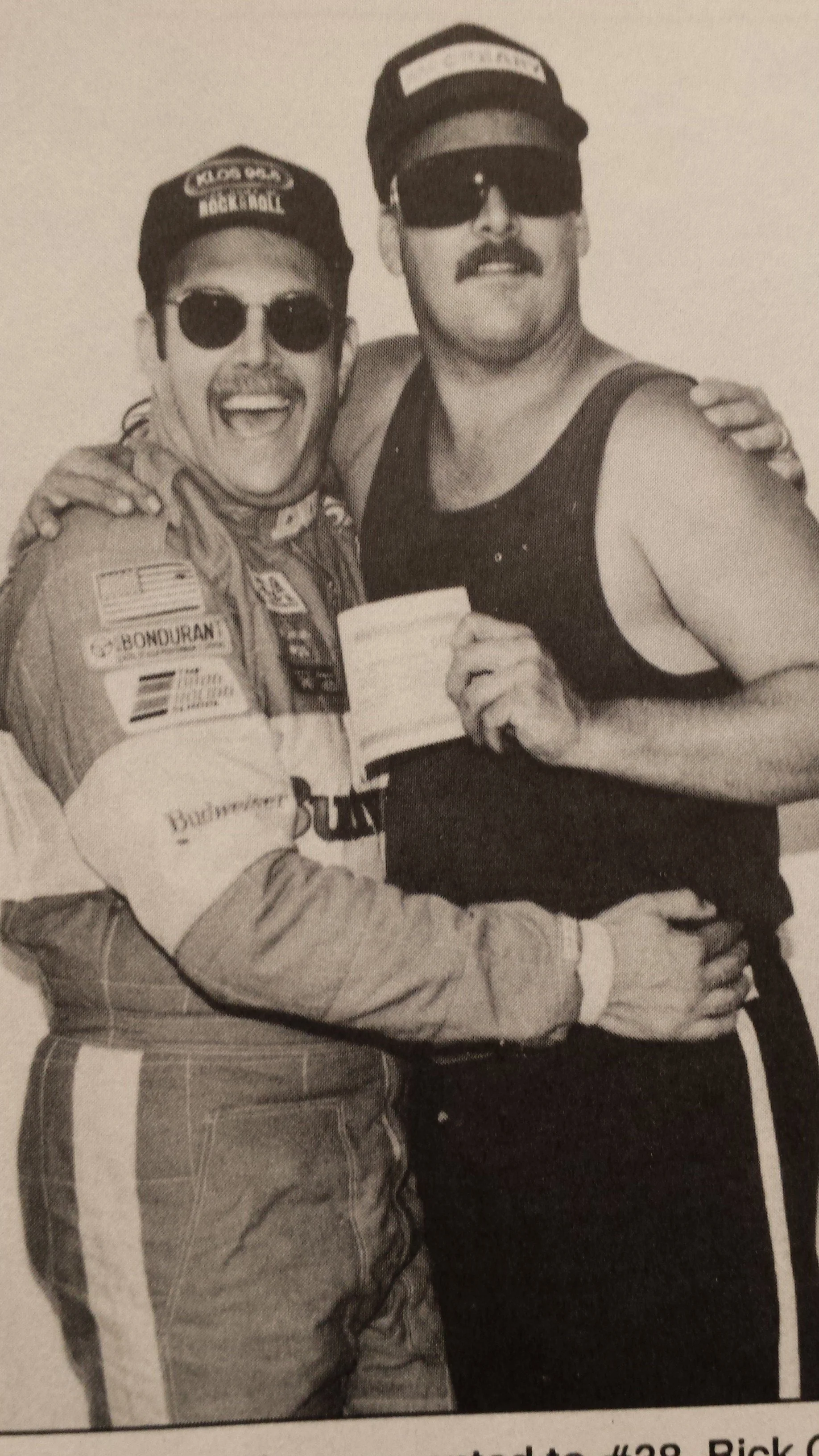 Black-and-white photo of two smiling people wearing caps and sunglasses, with their arms around each other.