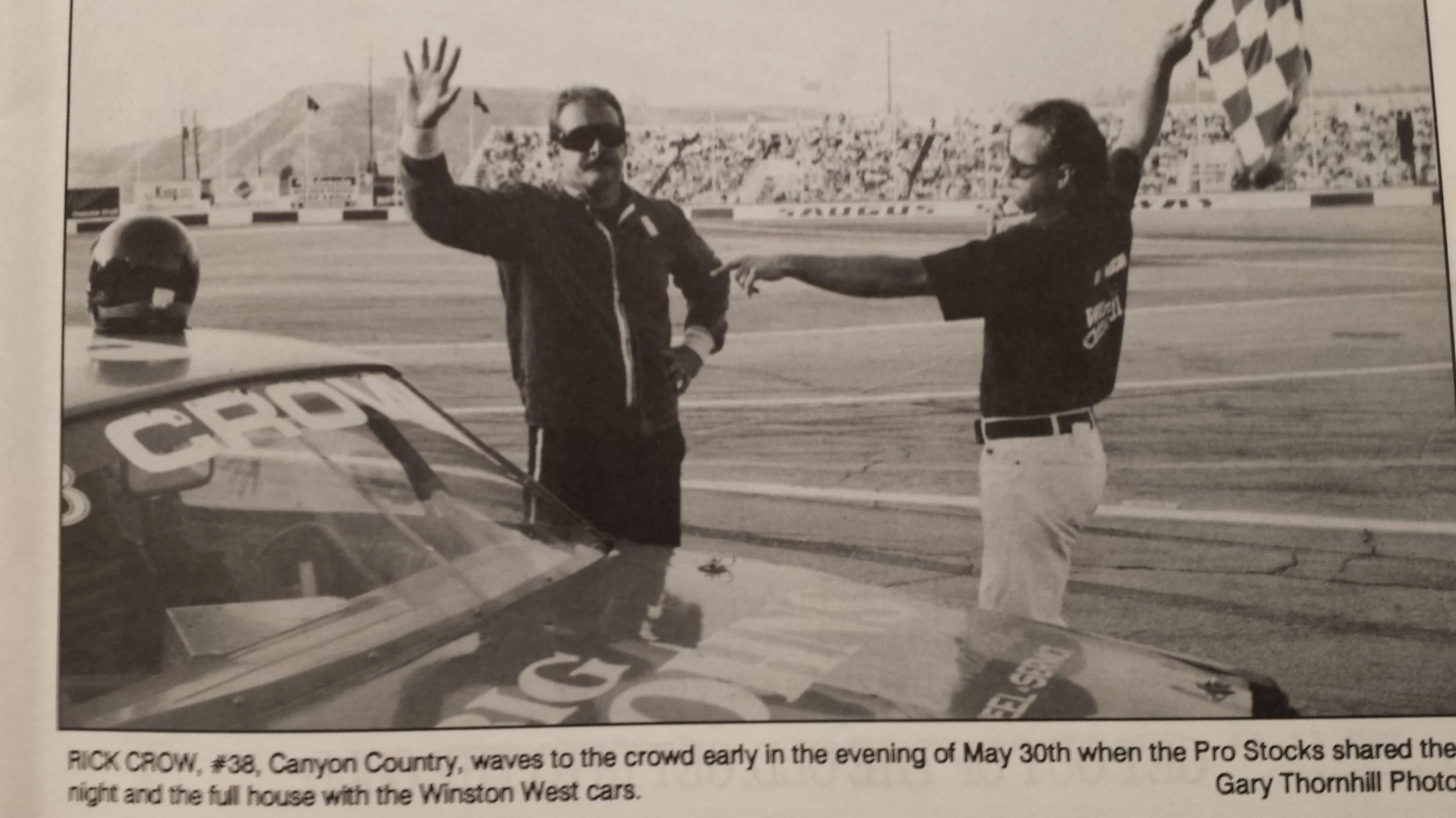 Black-and-white photo of race car driver Rick Crow waving beside a stock car on a track, with a crew member and checkered flag in the background.