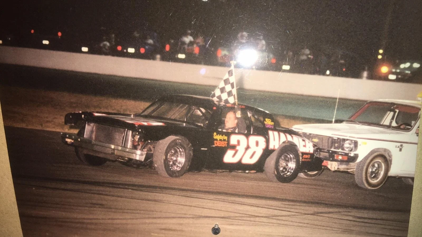Vintage black No. 38 stock car crossing the finish line with a checkered flag at a nighttime dirt track, alongside a white race car
