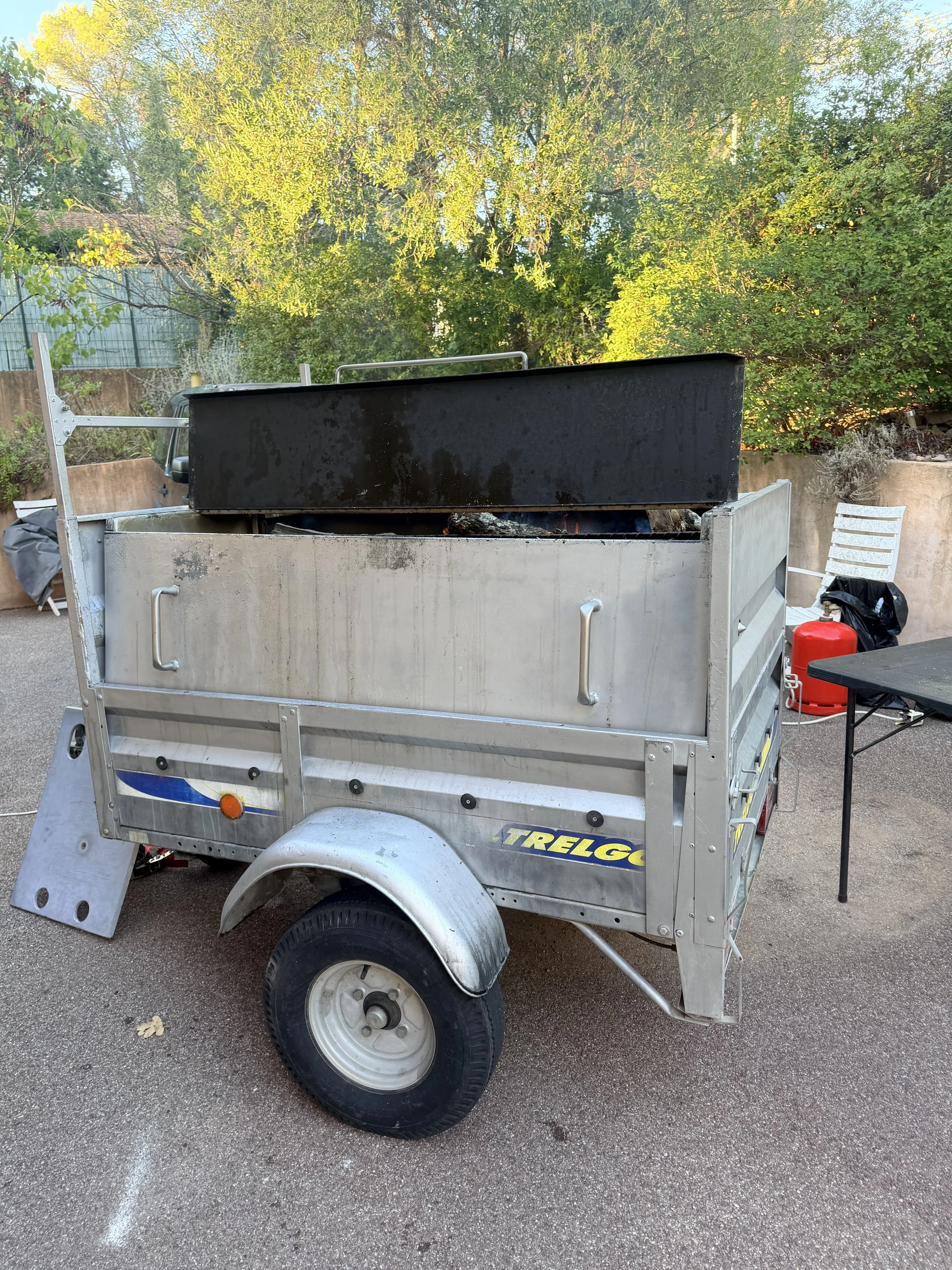 Remorque métallique contenant un grand barbecue à bois, installée dans un jardin arboré.