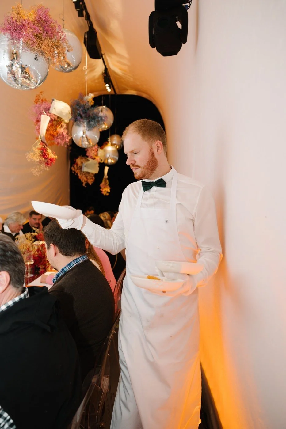 A waiter in a white apron serving dinner beneath hanging flower installations