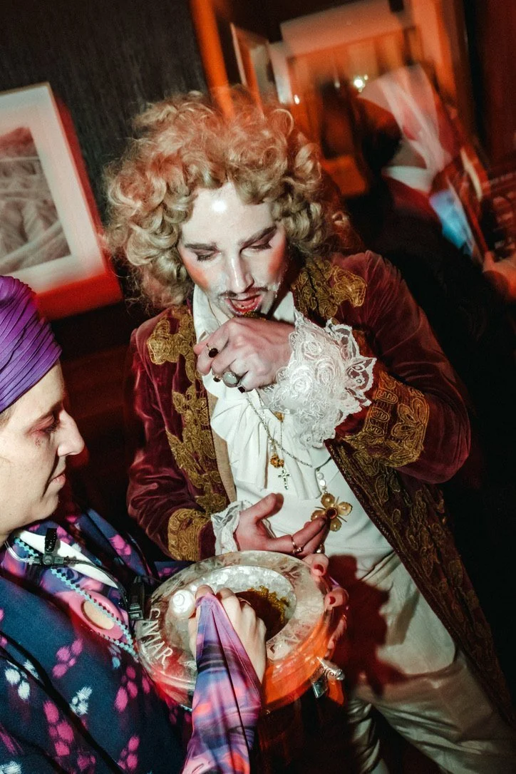 A guest in a baroque wig and velvet coat tasting caviar