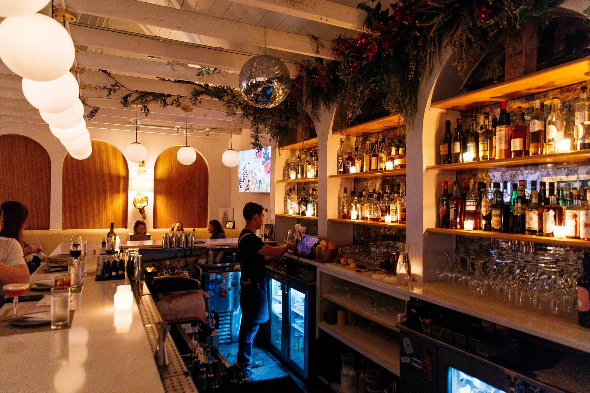 Hen House’s warmly lit bar with arched shelves, globe lights, greenery, and a disco ball