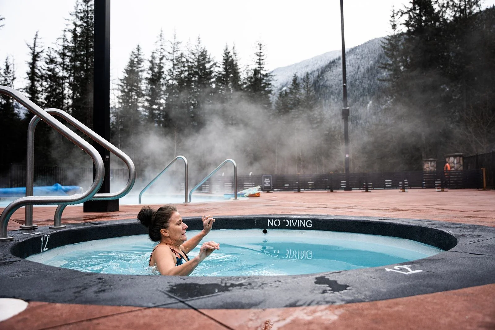 A woman easing into the cold plunge on a winter day, with steam drifting off the hot pools behind