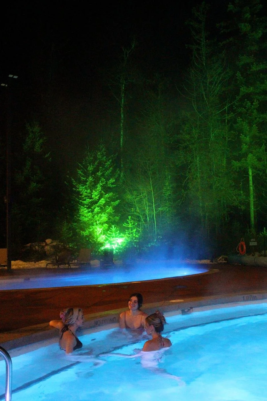 Three friends chatting in a hot pool at night, with forest lit green behind